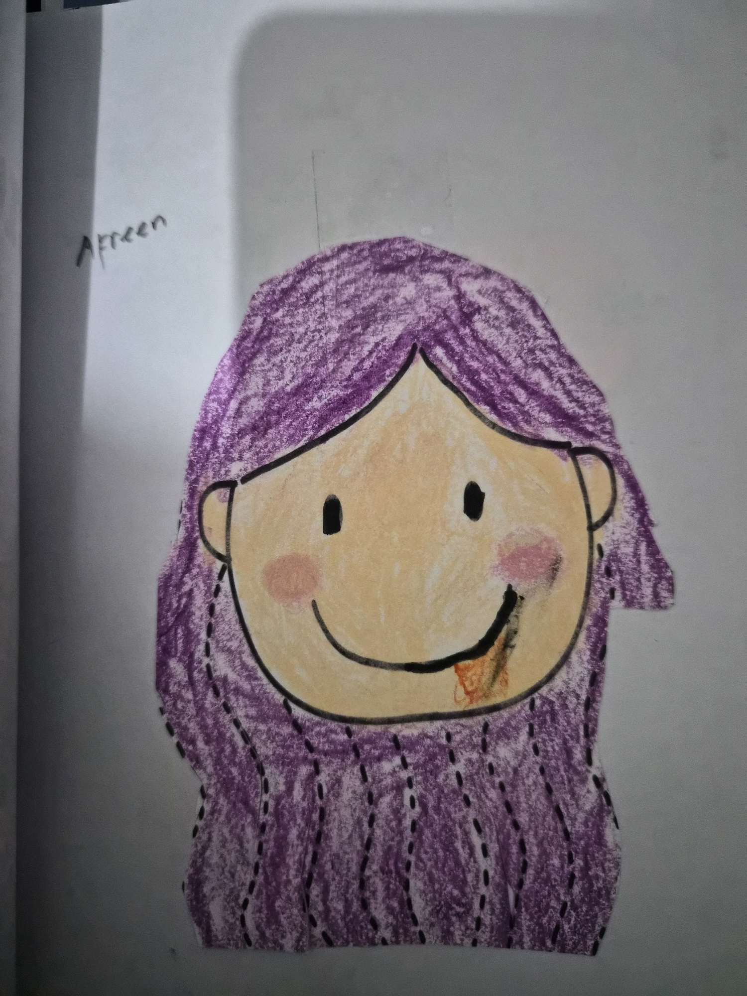 An overhead, slightly angled close-up shot features a drawing on white paper. The drawing is of a person's head and shoulders, rendered in crayon. The figure has purple hair, a light tan face with a black eye, a pink cheek, and a smiling mouth with orange color within. A name, "Afreen", is written in black marker in the upper left corner of the image, just above the figure's hair. The background is a plain, light gray. There are no discernible elements in the image that suggest a location in the city of Balikpapan, Indonesia.