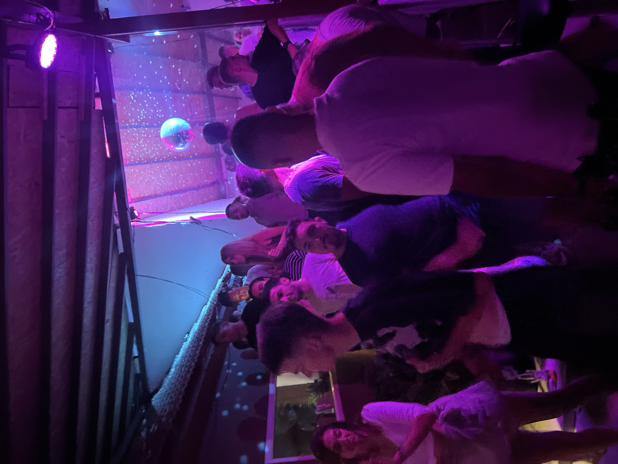 The image depicts an indoor scene at a crowded nightclub or bar, illuminated by purple and pink lights. A disco ball hangs from the ceiling, reflecting light across the space. Several individuals, mostly young adults, are visible, some dancing and socializing. The overall atmosphere suggests a lively evening event. While the location is described as being in Herceg Novi, Montenegro, this specific detail cannot be visually confirmed from the provided image alone.