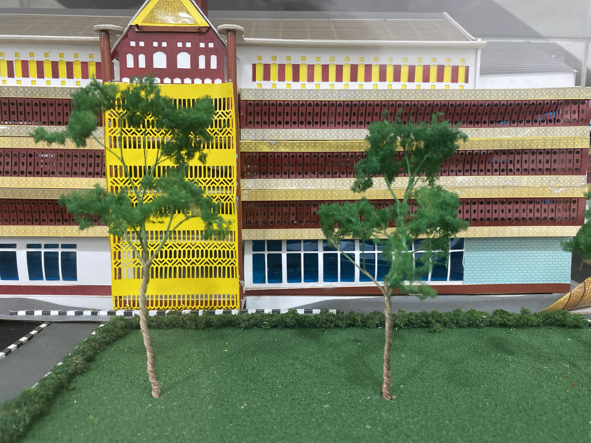 This is a photograph of a detailed architectural model of a building, possibly a school or public institution, in Banda Aceh, Indonesia. The model features a multi-story structure with a distinctive facade. The building's exterior is a vibrant combination of colors, including red, yellow, white, and gold. It exhibits repetitive patterns and decorative elements. The model includes realistic green trees with twisted brown trunks, placed in front of the building. A patch of green artificial grass represents a lawn, bordered by a low hedge. A section of a road with black and white pavement markings is visible on the left side of the image. The overall impression is of a well-crafted miniature representation of a building and its immediate surroundings.