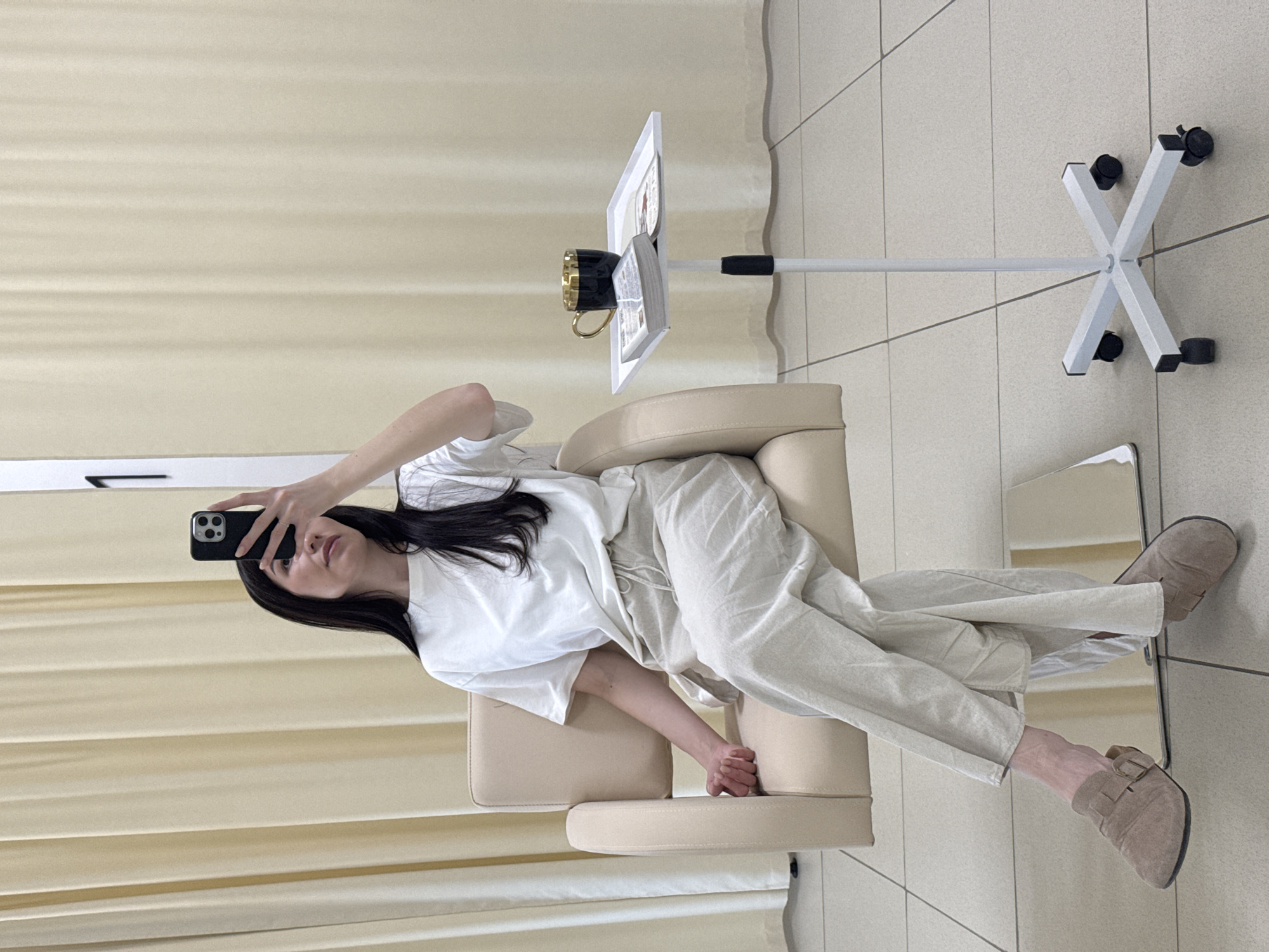 A single adult female is seated in a light beige, upholstered salon-style chair within an indoor setting. She has long, dark hair and is wearing a white short-sleeved t-shirt, light beige drawstring pants, and tan slip-on shoes with a buckle detail. Her right hand holds a black smartphone, which she is directing towards herself, suggesting a selfie or video capture. Her left arm rests on the chair's armrest, and her ankles are crossed. To her right, a white mobile stand with a rectangular tray supports a black mug with a gold interior and an open book or magazine. The background consists of light beige vertical curtains, and a white wall section with a large black number "1" adhered to it. The floor is tiled with light-colored, square tiles. The specific location within the city of Chaikovsky, Russia, cannot be determined from the visual evidence.