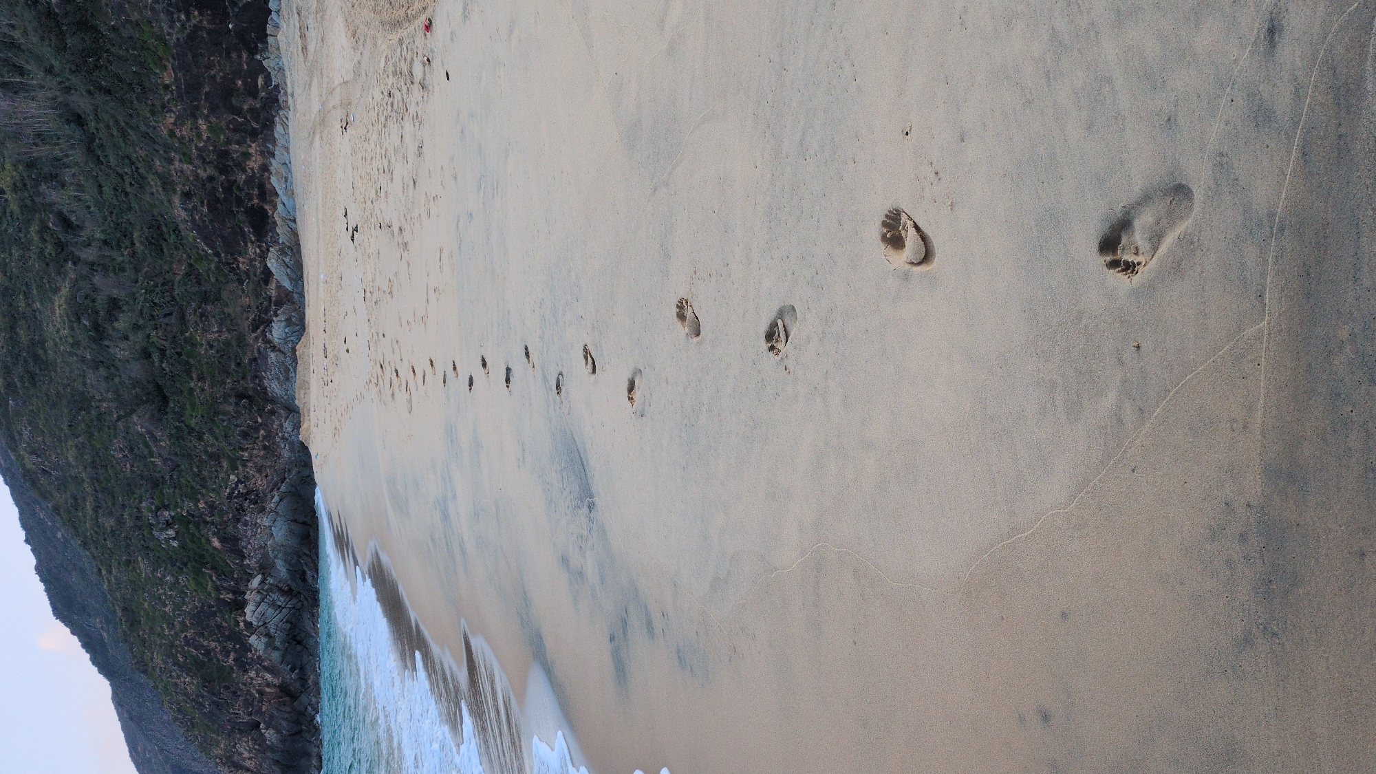 The scene depicts a sandy beach in East Quy Nhon Ward, Vietnam. A single file of approximately 14 human bare footprints is impressed into the light-colored, damp sand, leading from the lower right foreground diagonally towards the center-right of the background. To the left of the footprints, the ocean water is visible, characterized by a turquoise hue and white foam from gentle waves receding from the shore. The waterline shows the transition between wet and drier sand. In the background, a steep, dark green hill or mountain, partially covered with sparse vegetation and rocky outcrops, rises directly from the coastline. The sky is visible as a muted light blue-grey above the mountain. No individuals are present in the visible frame.