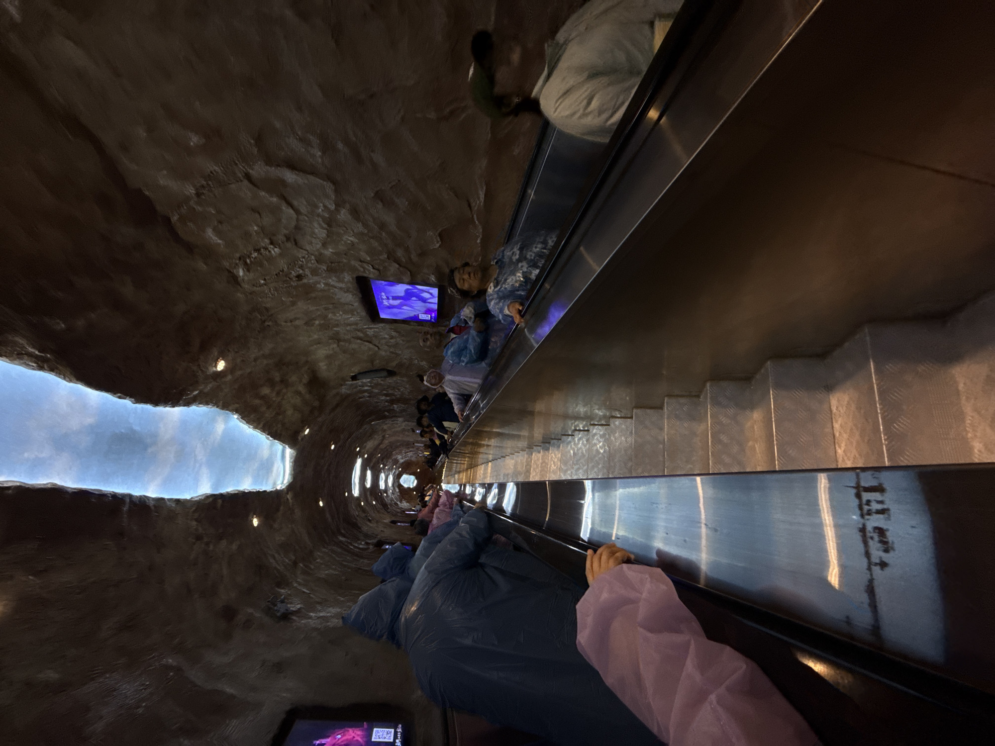 A high-angle shot shows an escalator inside what appears to be a large tunnel-like structure with a textured brown ceiling and a blue sky visible at the end of the tunnel. People are riding the escalator. Some individuals near the camera are wearing blue or pink clothing, possibly ponchos. A few television screens are mounted on the tunnel walls, displaying images. The location is in the city of Unknown, Unknown.