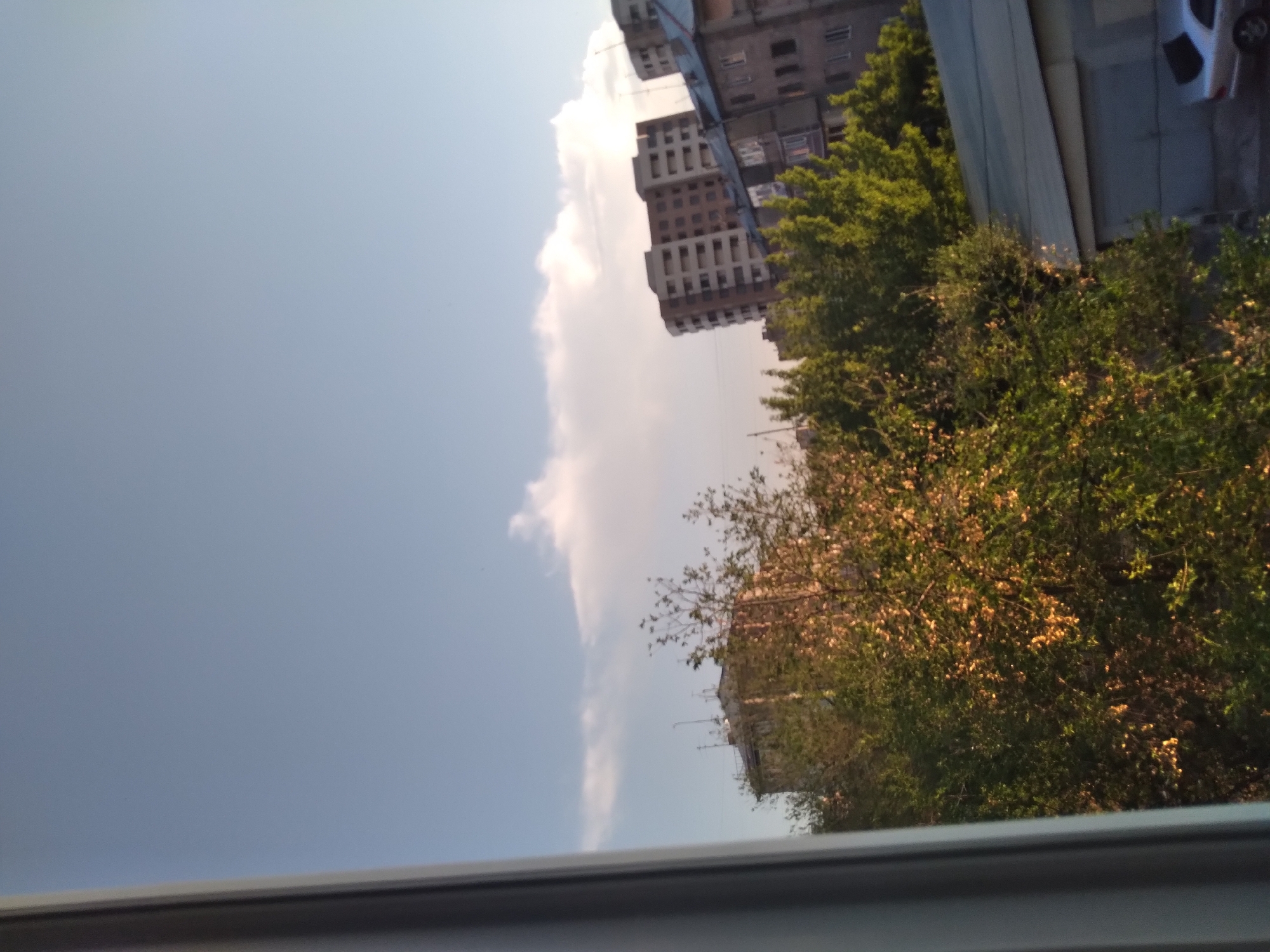 The image shows a view of an urban landscape with buildings and trees under a cloudy sky. In the foreground, a gray frame or windowsill is visible on the left side. Behind it, lush green trees are abundant, with some areas illuminated by sunlight, casting a warm glow on the foliage.
In the mid-ground, several apartment buildings are present. The building on the far right appears to be older and constructed with darker materials, possibly concrete or stone. It has multiple windows and a visible roof structure. To its left, a more modern, multi-story apartment building with a lighter facade and numerous balconies is depicted. This building is wider and has a slightly curved shape. Further to the left, another apartment building, appearing older and less detailed, is partially obscured by trees.
In the background, the sky is a clear blue with scattered white clouds. The lighting suggests it might be late afternoon or early morning, with the sun casting long shadows and highlighting certain parts of the scene. On the far right of the image, a silver car is parked in front of a garage or shed with a sloped roof. The scene is characteristic of a residential area in a city.