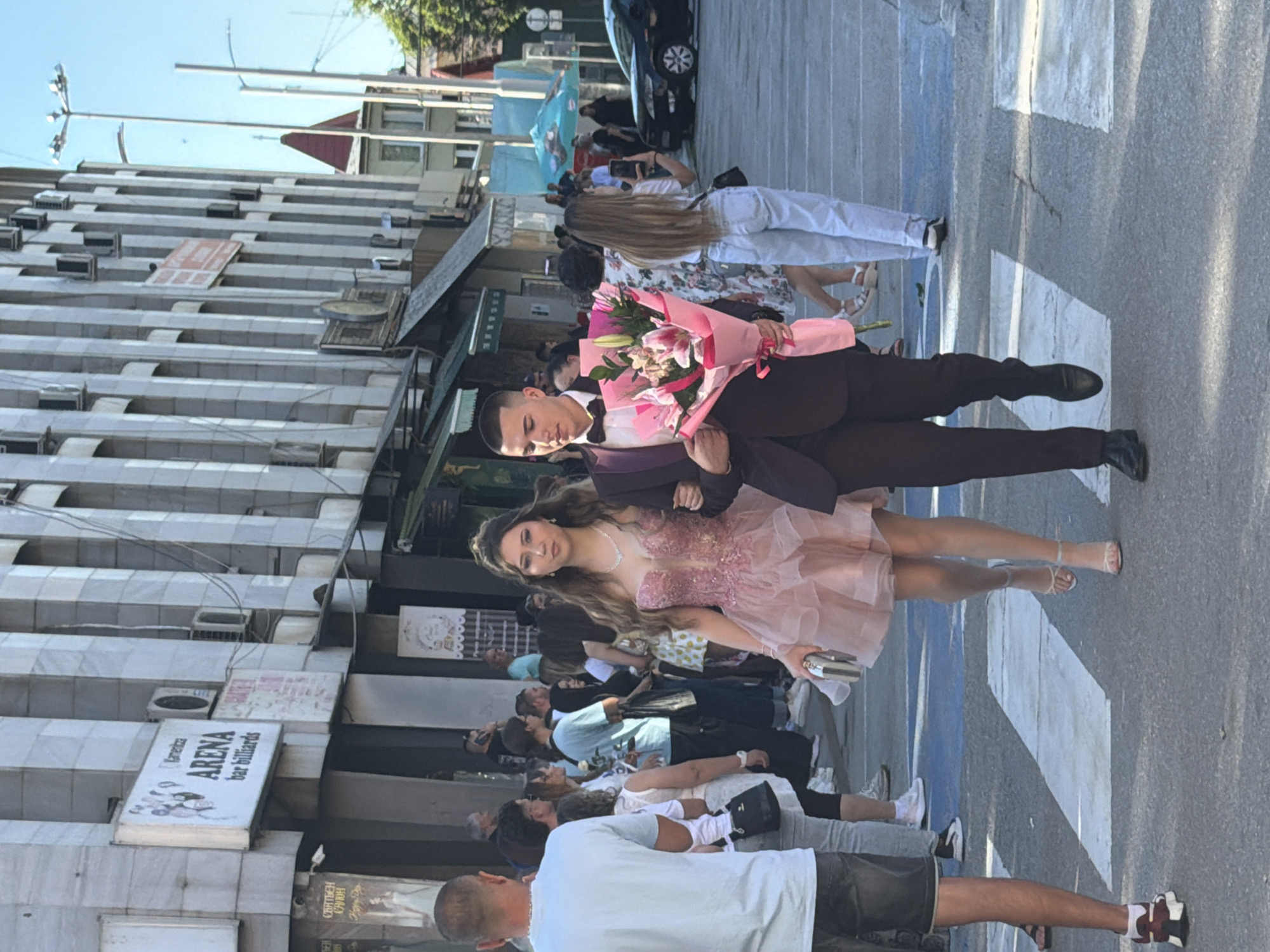 A vertically-oriented photograph captures a couple posing with a bouquet of flowers on a street in Pleven, Bulgaria. The woman is wearing a pink dress and the man a brown suit with a bow tie. They are centrally located in the foreground, with a crowd of people visible behind them. The background features a building with a prominent sign reading "ARENA bar billiards". The street itself has white painted markings. The overall atmosphere suggests a celebration or special occasion.