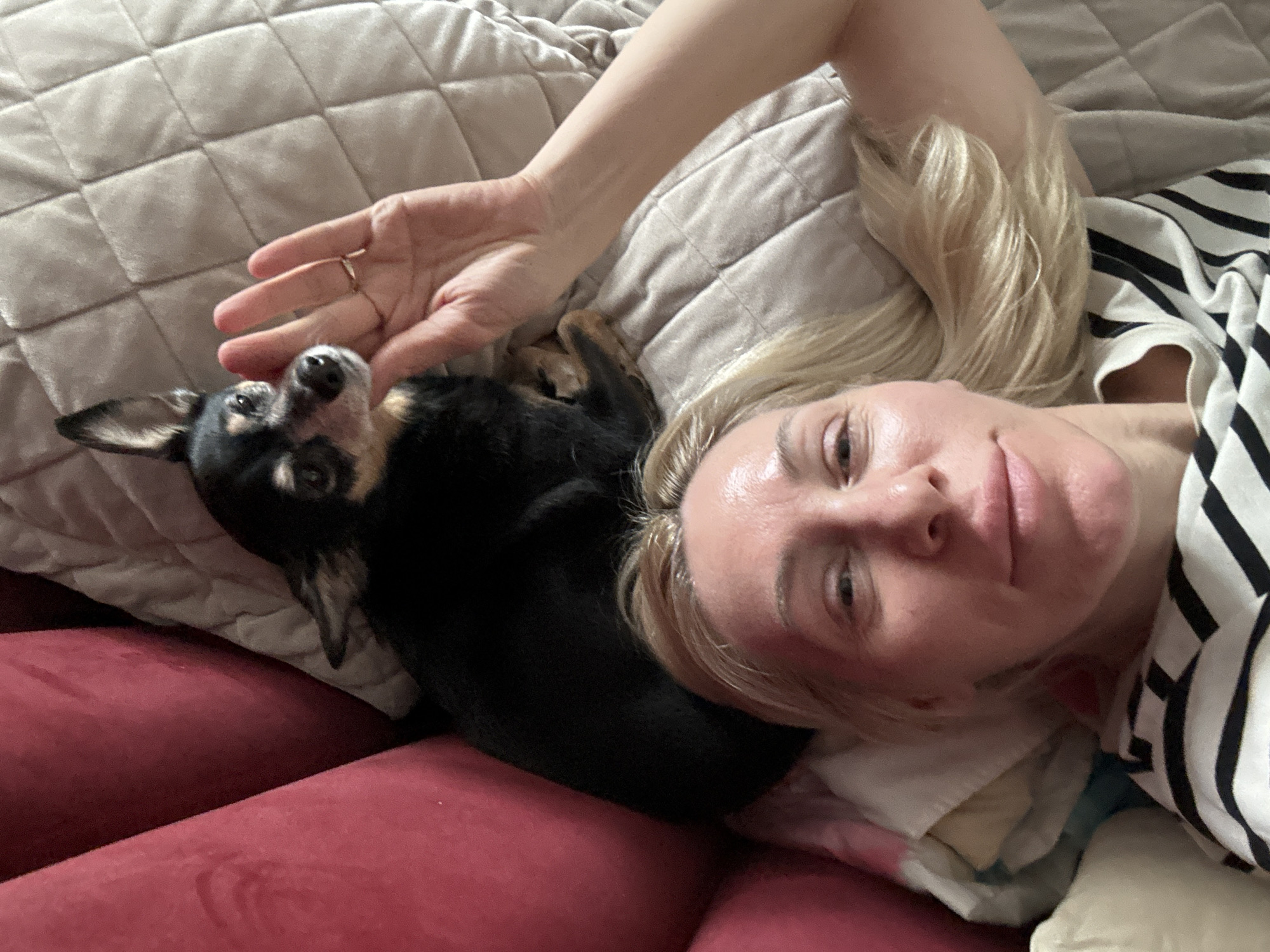 A woman with blonde hair is lying on a beige quilted pillow and red fabric. She is wearing a white and black striped shirt. To her left is a small black and tan dog. The woman's hand is near the dog. The location appears to be indoors.