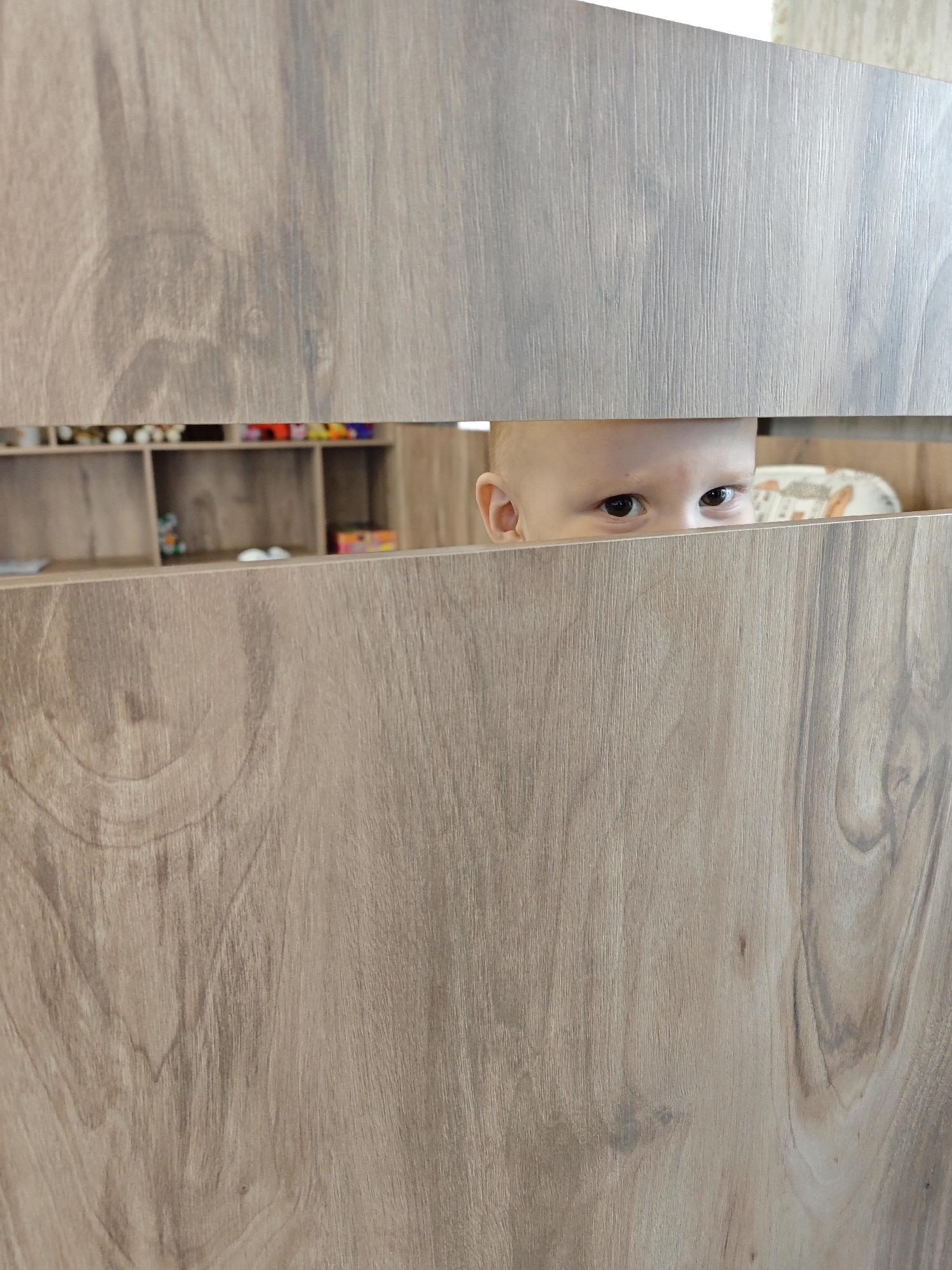 The image shows a close-up, eye-level perspective of a baby or toddler peeking out from behind a wooden barrier, presumably a crib or playpen, in an indoor setting in Chaikovsky, Russia. The foreground is dominated by two horizontal panels of light brown, wood-grained material, which obscure most of the child's body.
Through a narrow horizontal slit between these panels, only the upper part of the child's head is visible. Their alert, dark eyes are fixed directly at the viewer, conveying a sense of curiosity and engagement, while one ear is also clearly seen. The child's skin appears fair.
In the background, behind the child, a wooden shelving unit with several cubbies or compartments is partially visible. These shelves hold various small, colorful objects, likely toys, suggesting a child's room or a family living area. A patterned fabric or blanket is also discernable near the child.
The scene captures a playful moment, with the child appearing to be looking out or playing peek-a-boo. The lighting indicates it is daytime, and the overall atmosphere is domestic and intimate. There is no visible text in the image.