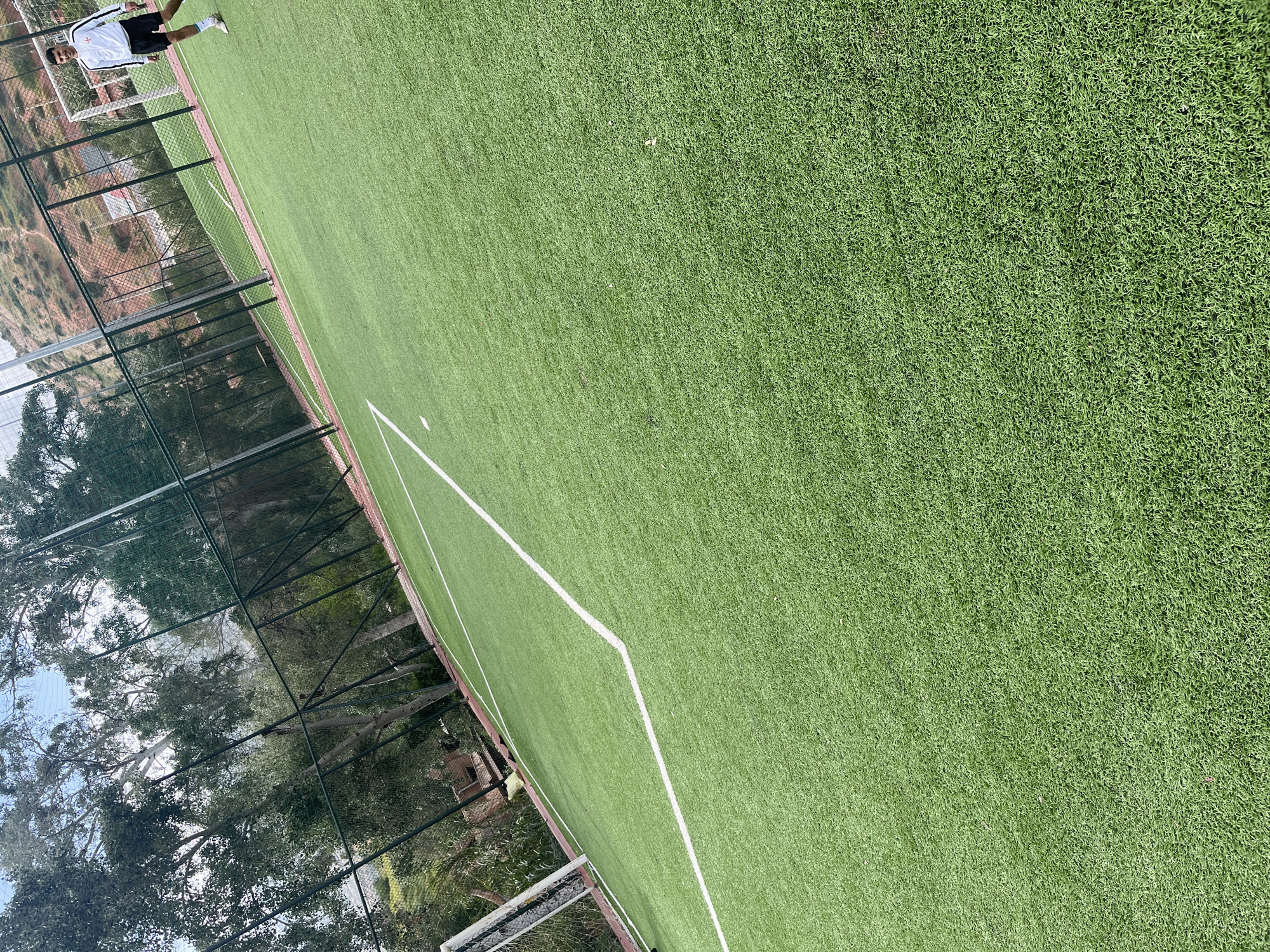 This image, captured from an elevated perspective and rotated approximately 90 degrees clockwise, depicts an artificial turf football (soccer) field located in Ouaouizert, Morocco. The lush green artificial grass dominates the foreground and midground, featuring clear white lines marking the field boundaries and a section of what appears to be a penalty area or central line.
In the upper left portion of the frame, a male individual is visible, standing near a white goal net. He is dressed in a white football jersey adorned with a red cross symbol (St George's Cross, associated with the English flag) on the chest, along with black shorts and light-colored sports shoes. He is looking towards the field. A second goal net is partially visible at the bottom right, suggesting the other end of the pitch.
The field is enclosed by a tall, dark green metal chain-link fence. Beyond this barrier, a natural landscape unfolds, featuring numerous mature trees with dense green foliage, particularly on the left and bottom edges of the image. Further in the background, a dry, hilly terrain with brownish-red earth is discernible, consistent with the provided location context of Morocco.
The scene is brightly lit by natural sunlight, indicating clear weather, likely during the late morning or early afternoon. While no active game or practice is taking place, the setup strongly suggests a venue for football activities. There is no visible text in the image.
