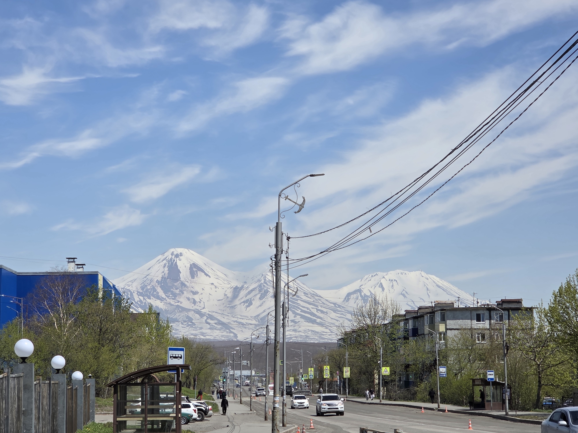 This image captures a wide street in an urban area with a striking natural backdrop under a bright, sunny sky. The location is Unknown, Unknown.
In the distant background, three prominent snow-capped mountains rise majestically against a light blue sky with scattered wispy white clouds. The mountain on the left appears to be the highest and most distinct. A lower, dark ridge of land, possibly forested, separates the mountains from the city.
The midground is dominated by a multi-lane asphalt road, likely a main thoroughfare. Numerous vehicles, including sedans and SUVs of various colors (white, dark, green), are visible on the road, some in motion, others parked along the sides. A white SUV is prominent in the right-hand lane, moving away from the viewer. Orange traffic cones are intermittently placed on the pavement beside the right lane.
Lining the street are various buildings and street infrastructure. On the left, a modern-looking building with a bright blue facade is partially visible behind trees. On the right, older, multi-story residential buildings with balconies and varied window configurations are present. Abundant green deciduous trees with fresh foliage line the sidewalks and obscure parts of the buildings, suggesting spring or early summer.
Multiple utility poles with complex overhead power lines crisscross the frame. Several streetlights are mounted on these poles; one notable streetlight pole on the right features a decorative anchor design above its light fixture. Bus stops, marked with blue signs featuring a white bus icon, are situated on both sides of the road, each with a functional shelter structure. Yellow triangular road signs indicating pedestrian crossings are also visible.
Several pedestrians are present on the sidewalks. On the left, a person in dark clothing walks away from the camera. Other individuals are seen near the bus stops and walking along the right sidewalk.
In the immediate foreground on the left, a wooden fence is topped with three spherical white lamps mounted on short posts. The foreground also shows a paved sidewalk and curb separating it from the road. The clear shadows cast by objects indicate bright daytime conditions.