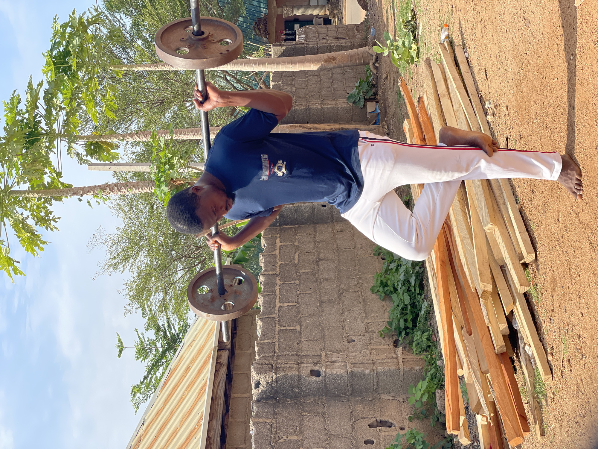 A man is lifting a barbell in an outdoor setting in Akwanga, Nigeria. He is wearing a dark blue shirt and white pants with a red stripe. He is barefoot. The barbell has two rusted metal plates. Behind him is a building with a brick wall and a corrugated metal roof. To his right are a stack of wooden planks on a dirt ground. In the background are trees under a partly cloudy sky.