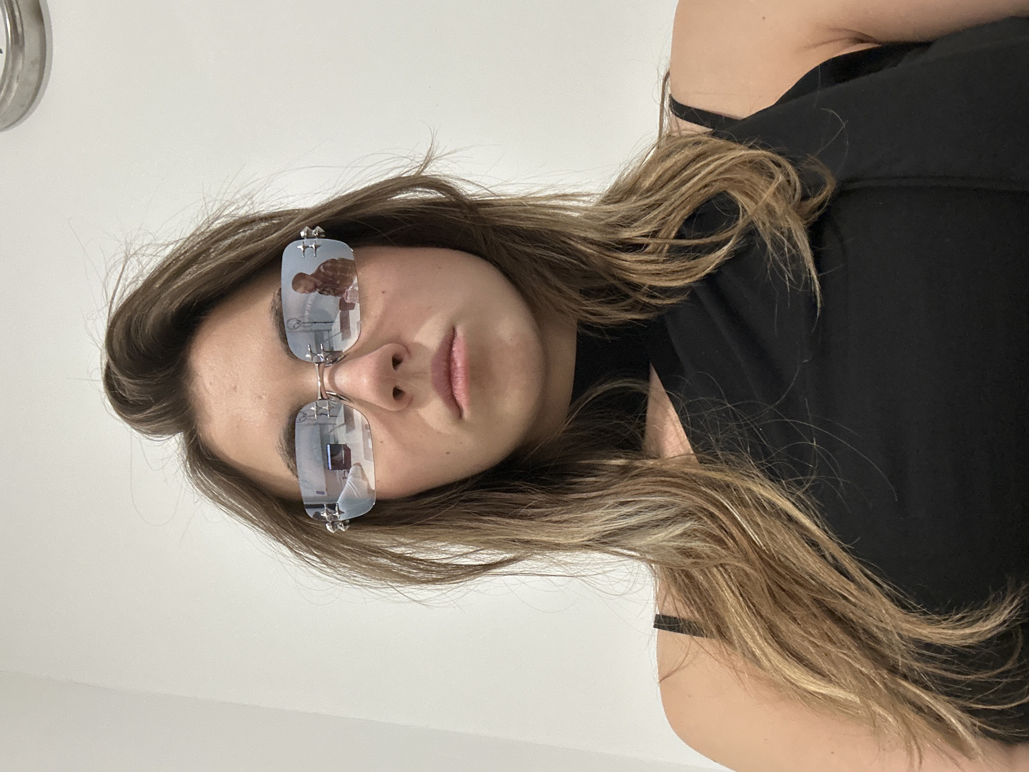 The image is a selfie of a young woman with long, wavy brown hair, wearing sunglasses and a black top. The sunglasses reflect a man wearing a plaid shirt. The background is a plain white wall. There is no indication of the location being in Moscow, Russia, or any other specific city.