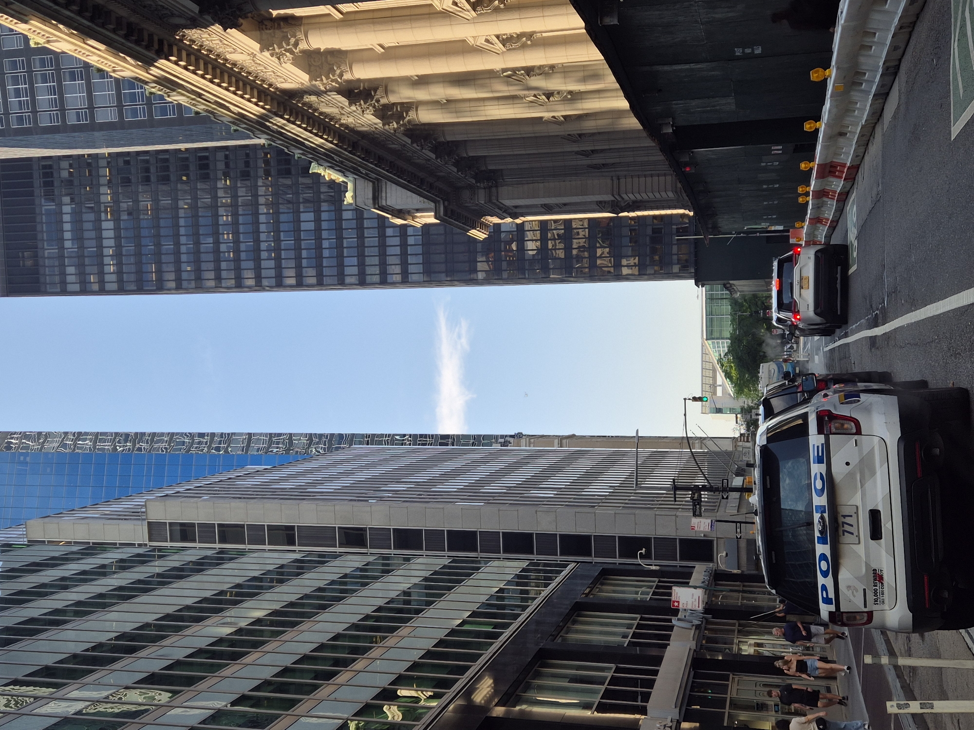 This is a street-level, eye-level, medium shot captured on a bright, sunny day in New York City, United States of America. The perspective is looking down a street lined with tall buildings. In the foreground, a white Ford police SUV is parked, with the word "POLICE" in blue lettering on the rear door and the number "771" on a license plate. To the left of the police vehicle, there are several people walking on the sidewalk, and further left, a glass-fronted building with many windows. To the right of the police vehicle, two lanes of traffic are visible, with a white SUV in the right lane. A temporary construction barrier with red and white sections and yellow lights lines the right side of the street, separating the traffic from a dark green wall, possibly a construction hoarding. In the background, more tall office buildings dominate the skyline, with a clear blue sky overhead. A traffic light is visible in the distance, showing a green signal. The overall scene depicts a busy urban environment.