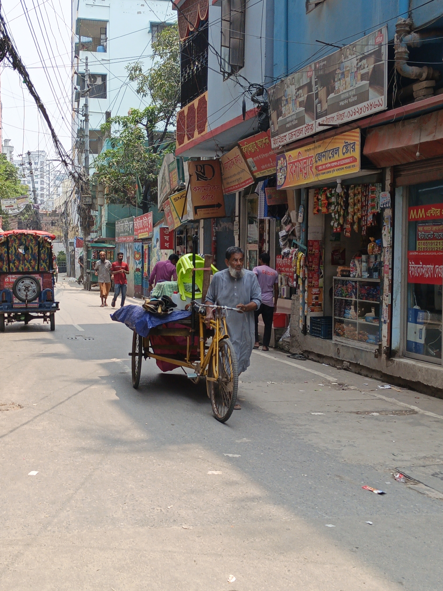 Here is a detailed description of the image:
The image shows a street scene in what appears to be a busy, older urban area in Bangladesh, potentially Dhaka based on architectural styles and signage. The street is relatively narrow and filled with activity. An older man with a grey beard is walking along the road, pushing a yellow cycle rickshaw laden with items covered by a blue tarp and a green chair.
Along the right side of the street are various small shops. A prominent shop front has signage in Bengali and English, advertising products and services including internet connections. Other shops sell assorted goods like snacks, drinks, and household items.
Other people are visible in the background: pedestrians walking along the street, a man in a red shirt, and rickshaw pullers. Buildings on both sides of the street show a mix of architectural styles, with visible electrical wires and signage adding to the cluttered visual environment. There's scattered litter on the street surface.
The overall impression is a lively, somewhat chaotic, yet normal day in a densely populated urban neighborhood.