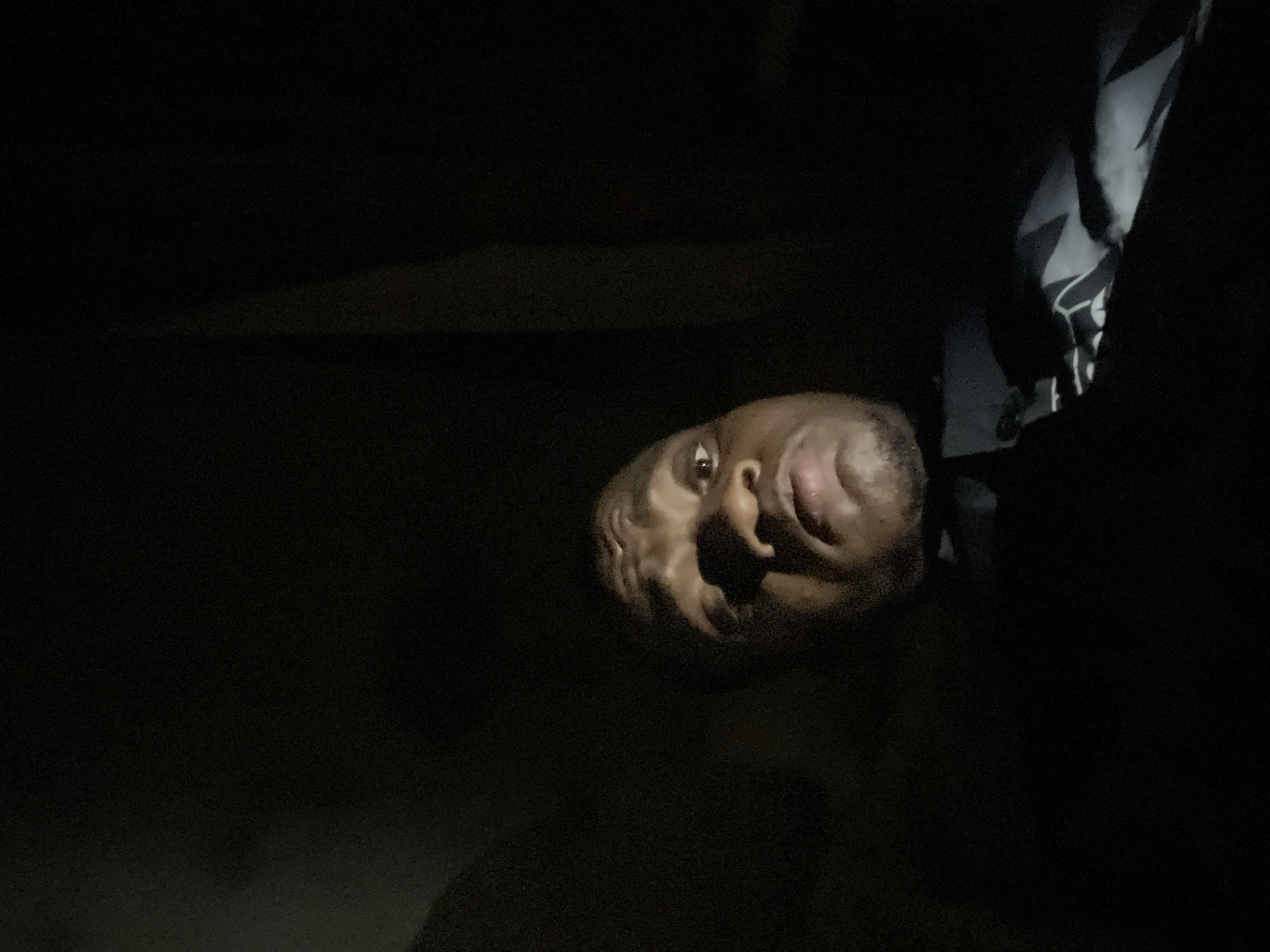 A photograph taken in low light depicts a person with dark skin and short hair, facing the camera with their mouth open, and appearing to be upside down in the frame. The background is dark, obscuring the environment. Some fabric is visible to the upper right of the person. There is no evidence to confirm that this image was taken in Minna, Nigeria.