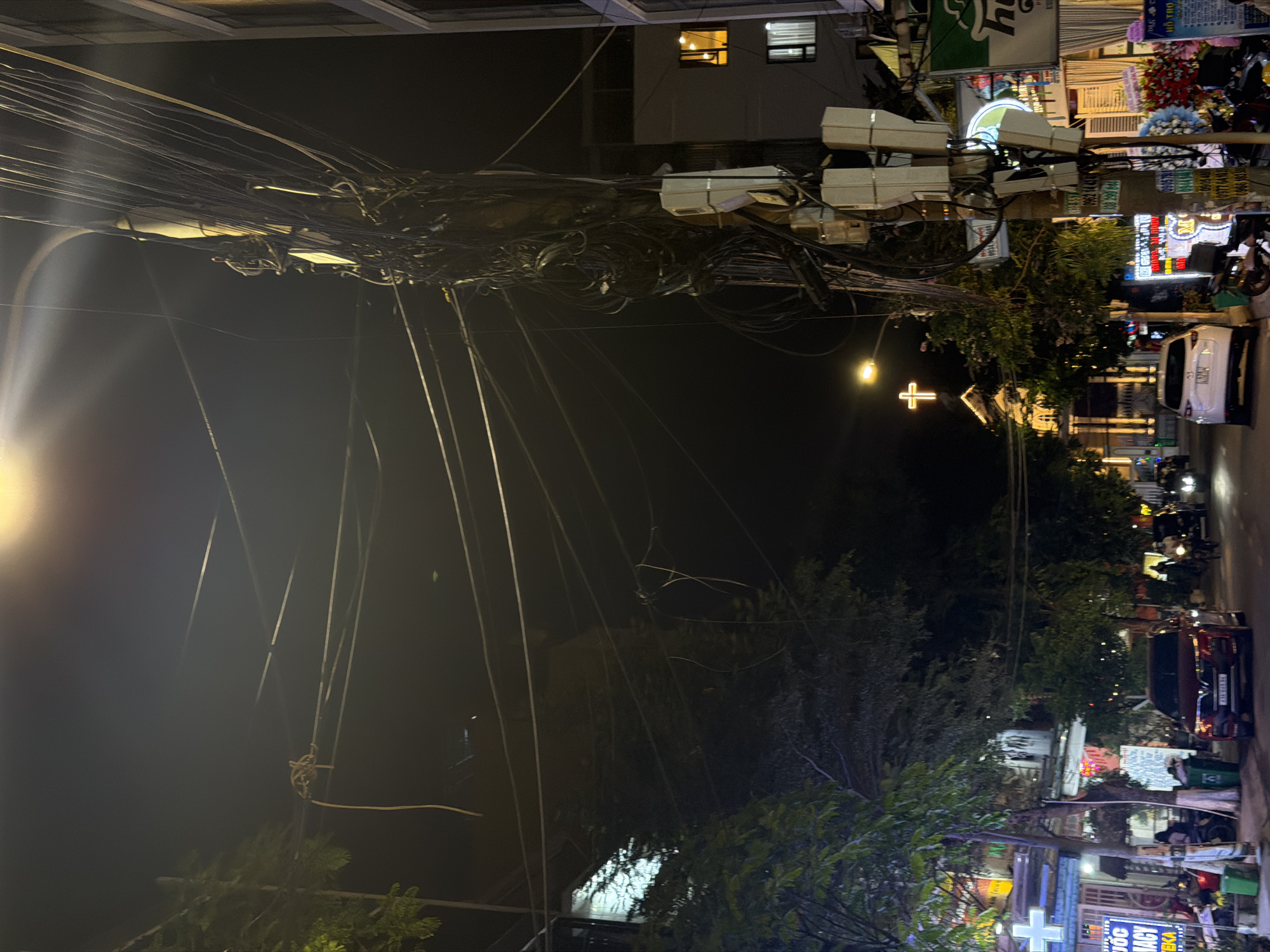This nighttime street scene in Đà Nẵng, Vietnam, is dominated by a chaotic network of overhead wires. The wires are tangled and dense, obscuring the view of the buildings and sky behind them. A Christian church with an illuminated cross is visible in the distance. Along the right side of the image, there is a row of shops with signs in Vietnamese. Parked cars and motorcycles line the street, and there are some people walking along the sidewalk. The scene is lit by streetlights and the lights from the shops.