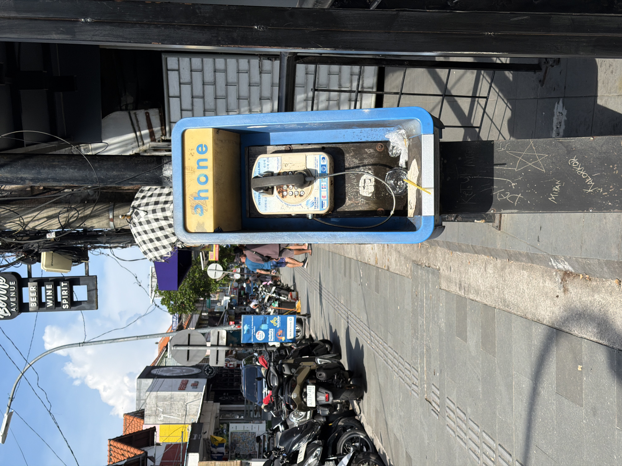 The image captures a weathered outdoor public payphone booth on a street in Kuta, Indonesia. The booth is blue with a yellow side panel displaying the word "phone." Inside the booth, the payphone is mounted, and a straw is placed in a glass of water next to it. The booth is attached to a black post with graffiti markings, including names like "MIRAN" and "KALORKOR."
In the background, the street scene reveals a busy area with numerous parked motorcycles. A sign for "Borre AVENUE BEER WINE SPIRIT" is visible, indicating the presence of a bar or similar establishment. A street lamp extends overhead, and other signs are also visible in the distance. Pedestrians are walking on the sidewalk, creating a sense of activity. The weather appears sunny, casting shadows on the ground.
