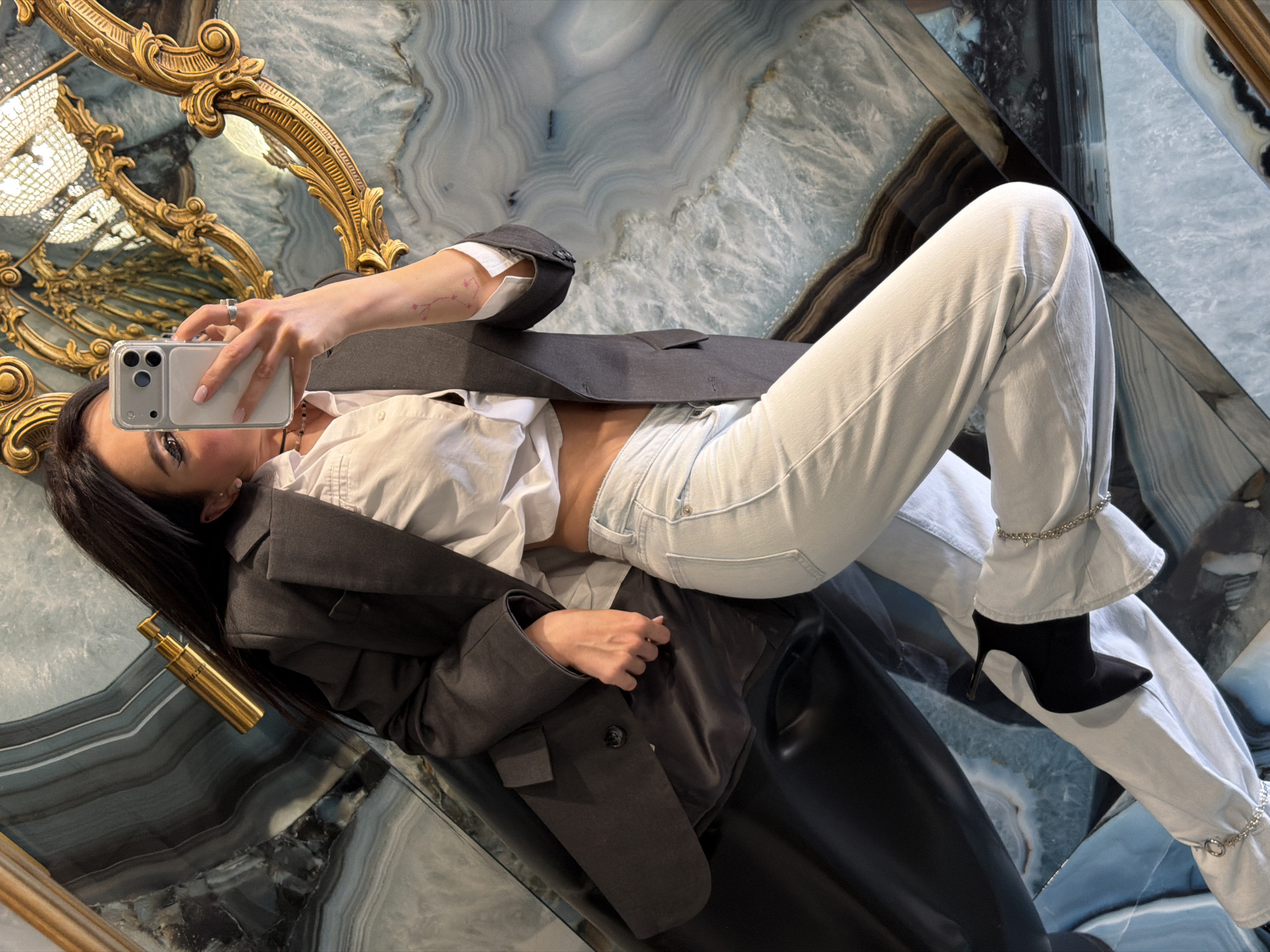 A person is depicted taking a mirror selfie, posing in a reclined position against a highly reflective surface that presents a striking, marble-like or agate pattern in shades of blue, gray, white, and brown. The individual is dressed in light blue jeans with unique chain details on the flared cuffs, paired with black high-heeled ankle boots. They wear a white button-up shirt, partially untucked to reveal the midriff, and a dark gray blazer. A silver ring is visible on one hand, and a small pink tattoo resembling a constellation is on their forearm.
The person holds a white/silver smartphone with a clear case, featuring prominent camera lenses, to capture the image. In the background, an ornate gold mirror frame is partially visible, reflecting what appears to be a chandelier. A gold cylindrical object, possibly a perfume bottle with faint, unreadable text, lies next to the person's head.
The overall scene suggests a luxurious and meticulously designed interior, characterized by its reflective surfaces and decorative elements. The image's tilted perspective adds a dynamic visual interest. This fashionable self-portrait is set in what is understood to be Moscow, Russia, consistent with the opulent aesthetic.