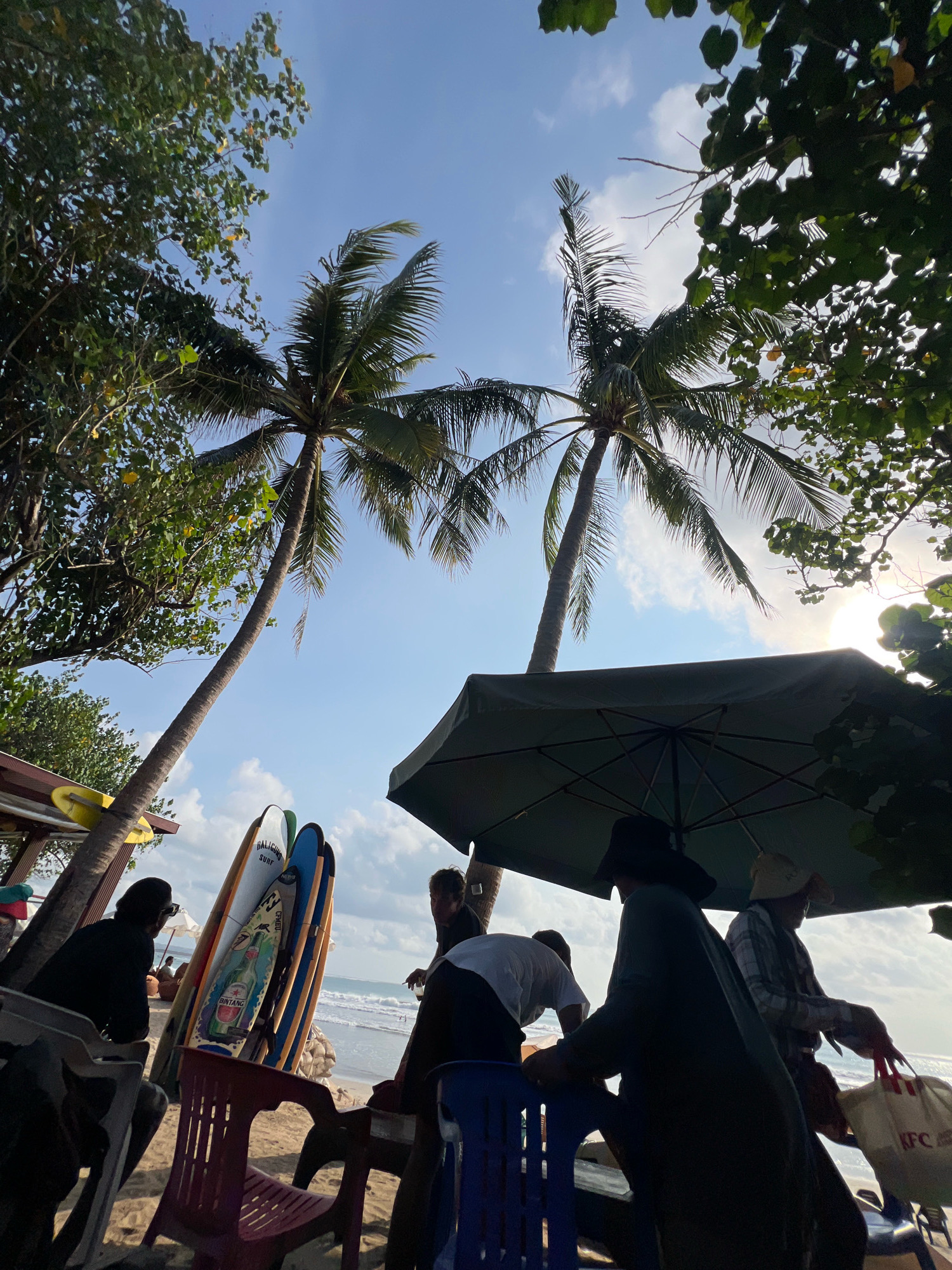 A low-angle, upward-facing shot captures a beach scene in Kuta, Indonesia, with two palm trees dominating the frame against a bright blue sky with scattered clouds. In the foreground, several people are seated at tables on the sand, some under a large, dark green umbrella. Beach chairs, including a red plastic chair and a blue plastic chair, are visible. A stack of surfboards is propped up on the sand, leaning against each other. The surfboards display various colorful designs and branding, including "BALIGUAN SURF" and what appears to be a "Bintang" beer advertisement. The ocean and beach are visible in the background. On the far right, a person holds a white bag with "KFC" logo. The overall atmosphere suggests a relaxed beachside gathering.