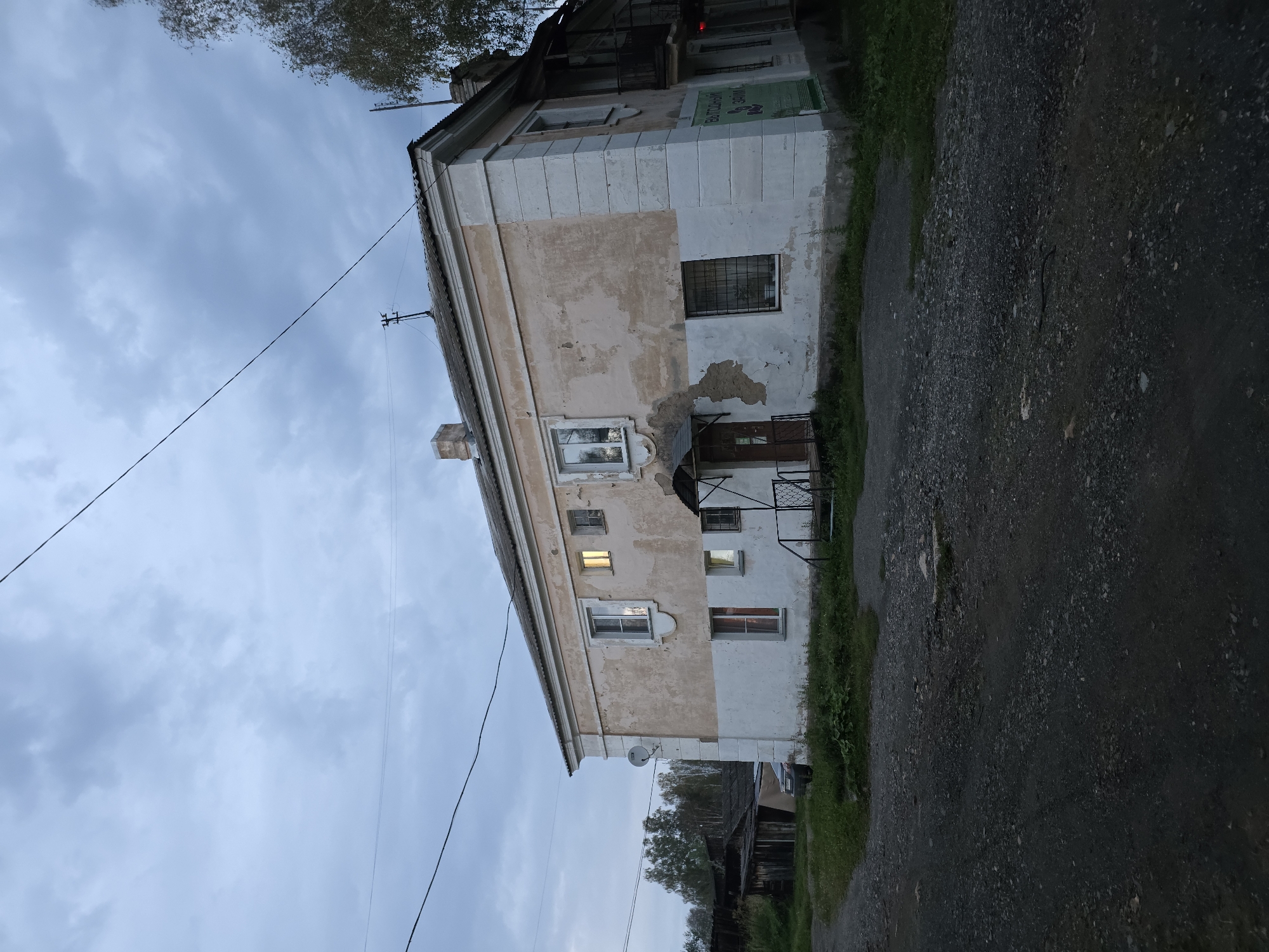 The image shows a two-story apartment building in Magnitka, Russia, under a cloudy sky. The building has a worn, stucco exterior with peeling paint, a mix of cream and white. There are several windows, some illuminated from within, and a small covered entrance with a dark door and metal railings leading up to it. A satellite dish is mounted on the side of the building. To the left of the building, there is a gravel driveway and a darker, weathered structure that appears to be a shed or outbuilding, with a car parked in front of it. Trees and foliage are visible in the background. A power line stretches across the upper portion of the image. The overall impression is of a somewhat dilapidated, older residential area.