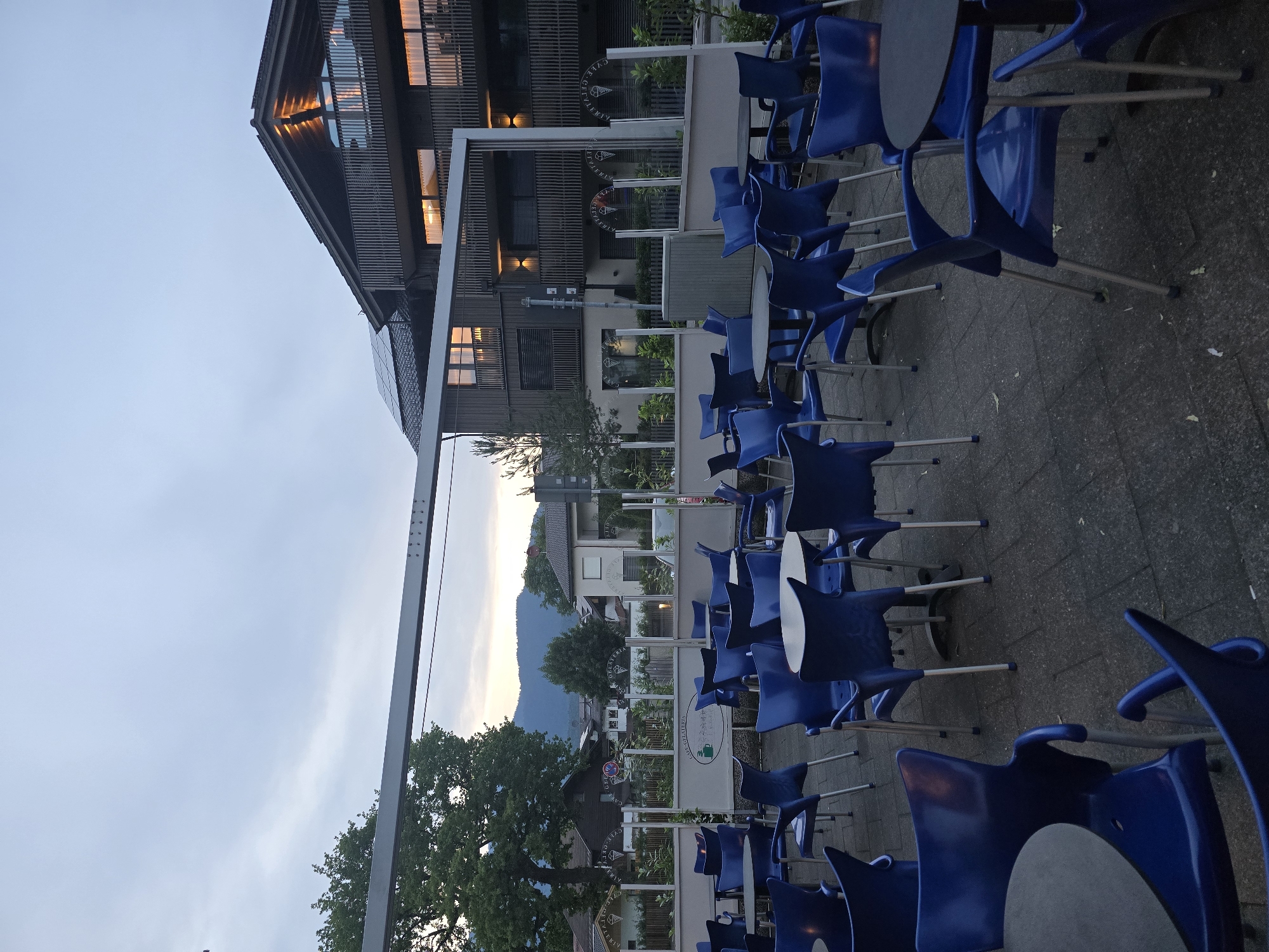The image depicts an outdoor seating area of a cafe or restaurant in Garmisch-Partenkirchen, Germany, as dusk settles. The scene is dominated by numerous blue plastic chairs arranged around dark-colored tables on a paved patio. In the background, a modern, multi-story building with dark wood siding and balconies is visible. The building features illuminated windows, suggesting indoor activity. Solar panels are present on the roof of this building. To the left of the building, a large, mature tree stands prominently. Further in the distance, houses and a tree-lined mountain range can be seen against a cloudy twilight sky. A white canopy structure extends over part of the seating area, supported by metal poles. A low barrier with signage that appears to be the cafe's name, "CAFE GELATERIA," runs along the back of the seating area. No people are present in the image.