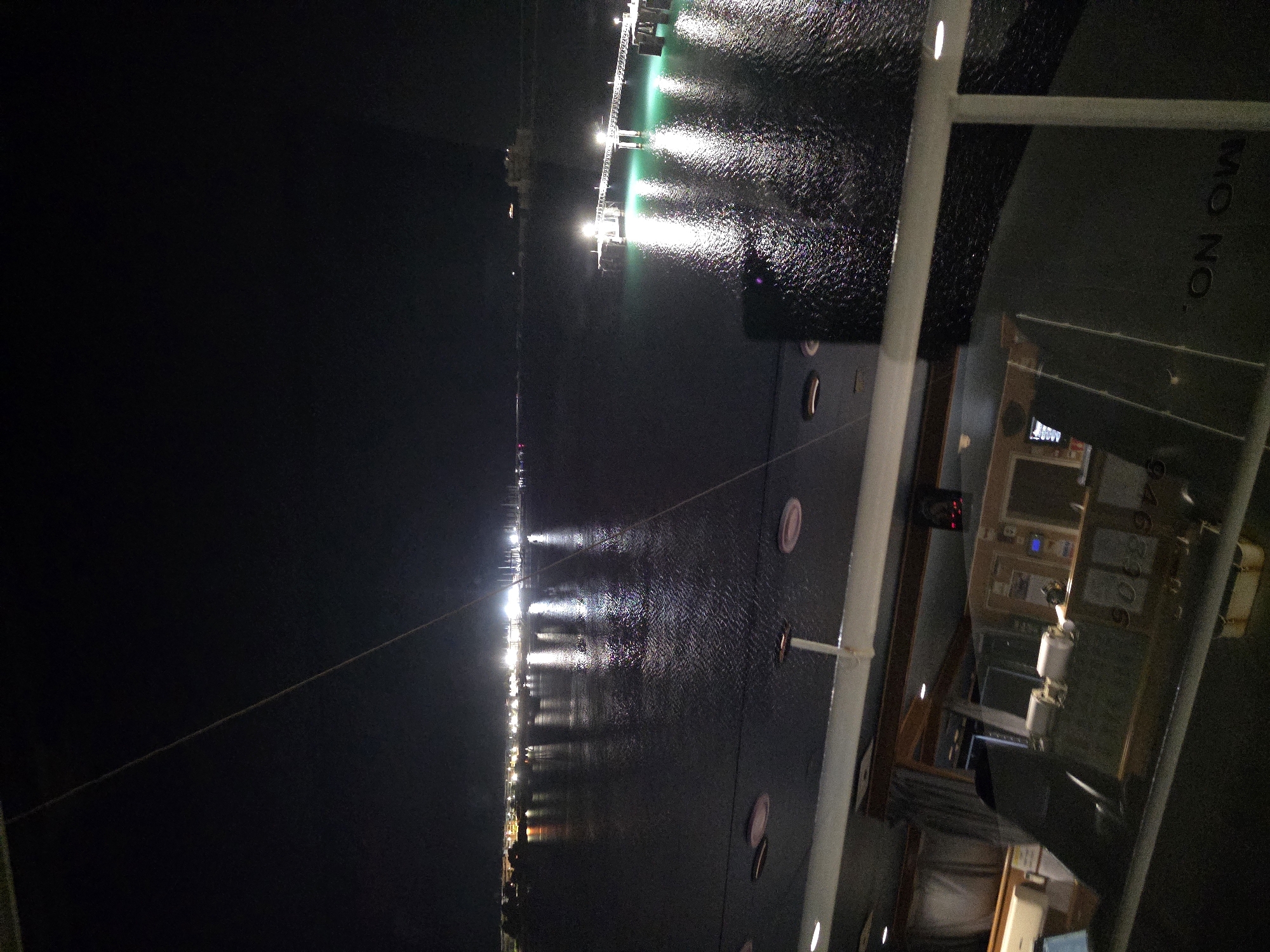 The image is a nighttime view of a waterfront in Brindisi, Italy, as seen from a vessel. The foreground shows a portion of the vessel's interior, including a control room with various instruments, consoles, and seating visible through a window. Numbers are visible on the control panels. The middle ground features a dark body of water, reflecting the lights from the city and a pier. In the background, a city skyline is illuminated with numerous lights, and a long pier with lights extends into the water. The water is dark and reflects the artificial lights, creating streaks of illumination. The sky is dark, indicating nighttime. The overall scene depicts an urban waterfront at night, with the perspective suggesting the viewer is on a ship or ferry.
