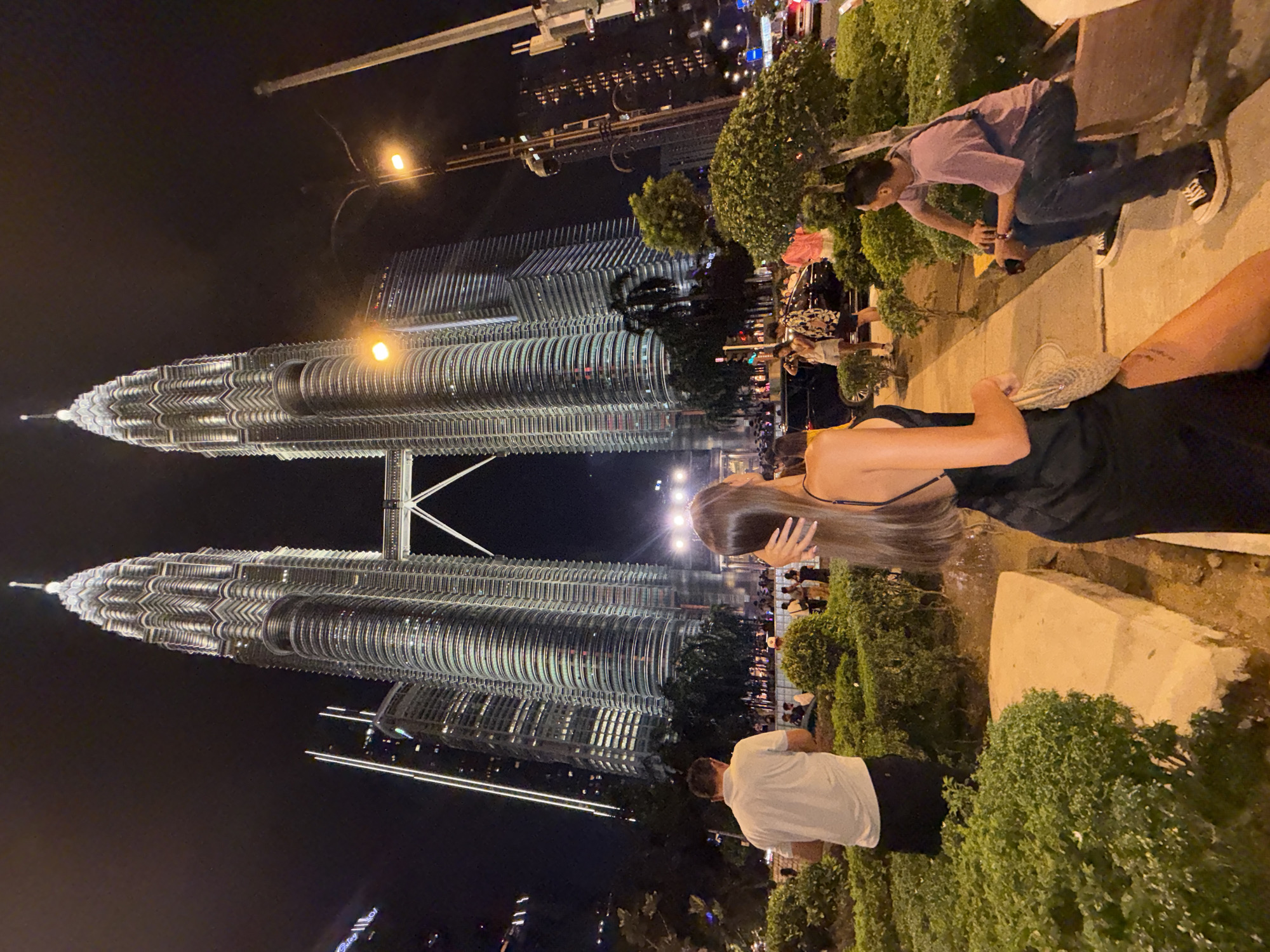 The image captures a night scene in Kuala Lumpur, Malaysia, with the Petronas Twin Towers illuminated prominently in the background. A crowd of people are gathered in the foreground. Several individuals are observed interacting with each other and their environment. The photograph showcases lush greenery along a sidewalk or pedestrian area, interspersed with concrete structures. Overhead street lighting and building lights contribute to the overall brightness of the scene.