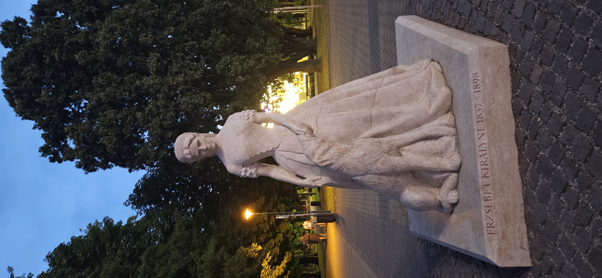 This is a medium shot, low-angle, full-frame photograph of a stone statue of Empress Elisabeth of Austria, known as Sisi, and her dog in Keszthely, Hungary. The statue is made of light-colored stone and depicts the empress in a voluminous, floor-length gown with off-the-shoulder sleeves. Her hair is styled up in an intricate updo. A dog sits attentively beside her, its head raised and looking towards her. The base of the statue is inscribed with the text "ERZSEBET KIRALYNE 1837 - 1898" in red lettering.
The statue is positioned outdoors on a paved walkway made of small, dark cobblestones. In the background, there are large, mature trees with dark green foliage. A lamppost with a warm orange glow illuminates the left side of the scene. Two individuals, a woman and a man, are walking away from the camera on the paved walkway in the mid-ground. The sky above is a deep blue, suggesting dusk or twilight. The overall ambiance is serene and commemorative. The statue is located in Keszthely, Hungary.