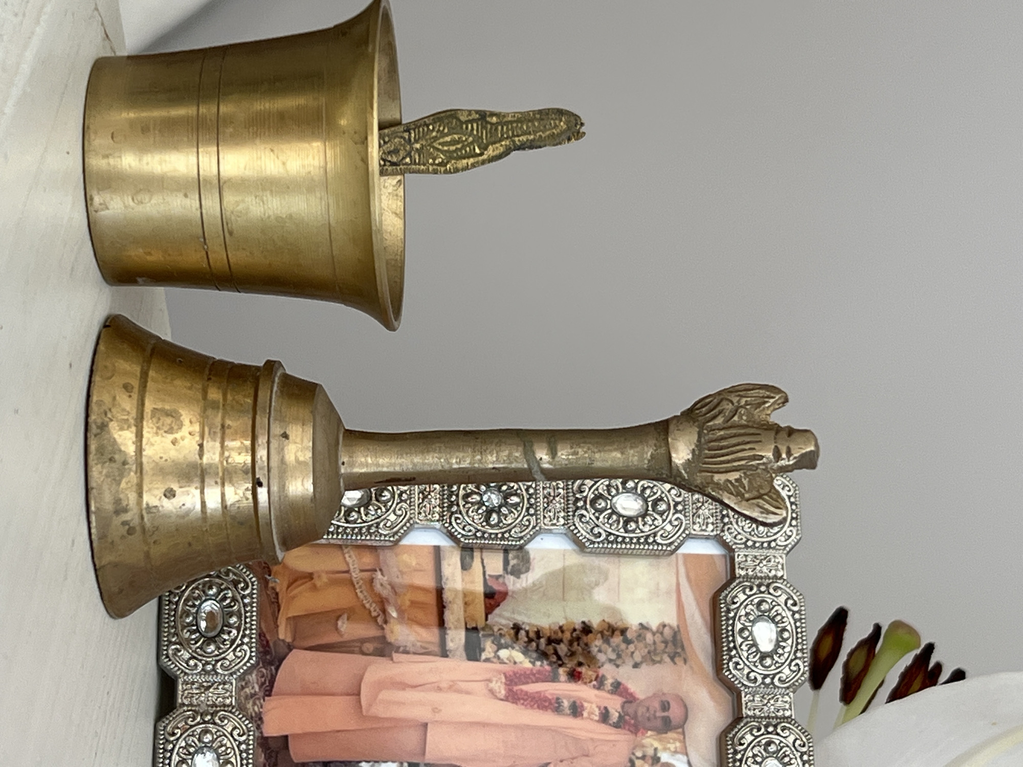 This media file displays two brass bells, likely used for religious or ceremonial purposes, positioned near a framed photograph. The top bell, shown from the side, is a simple, cylindrical shape with a flared opening. Inside this bell, a decorative element, possibly a clapper, is visible. It appears to be an ornate, stylized animal or serpent form, intricately carved with a pattern.
The second bell, positioned below and to the left of the first, has a more elaborate design. It features a handle topped with a decorative finial that resembles a claw or a stylized bird's foot. This bell is set against a framed photograph. The frame itself is ornate, with a silver-toned metal border decorated with intricate patterns and inset with small, clear gemstones.
The photograph within the frame shows a man dressed in peach-colored robes. He is adorned with a garland of flowers around his neck, suggesting a special occasion or religious ceremony. The man has a shaved head and appears to be lying down or seated, with his face visible. The background of the photograph is somewhat indistinct but seems to suggest a gathering or devotional setting, possibly with more floral decorations.
The lighting in the image is soft and appears to be from an indoor source, as there are no strong shadows indicative of direct sunlight. The overall impression is one of reverence and cultural significance, with the brass bells and the framed image suggesting a religious or spiritual context. The presence of white petals in the foreground, partially obscuring the frame, adds a touch of natural element to the scene, possibly hinting at a recent ritual or offering. The setting appears to be indoors, likely a home or a dedicated space for worship or remembrance. There is no visible text in the media file.