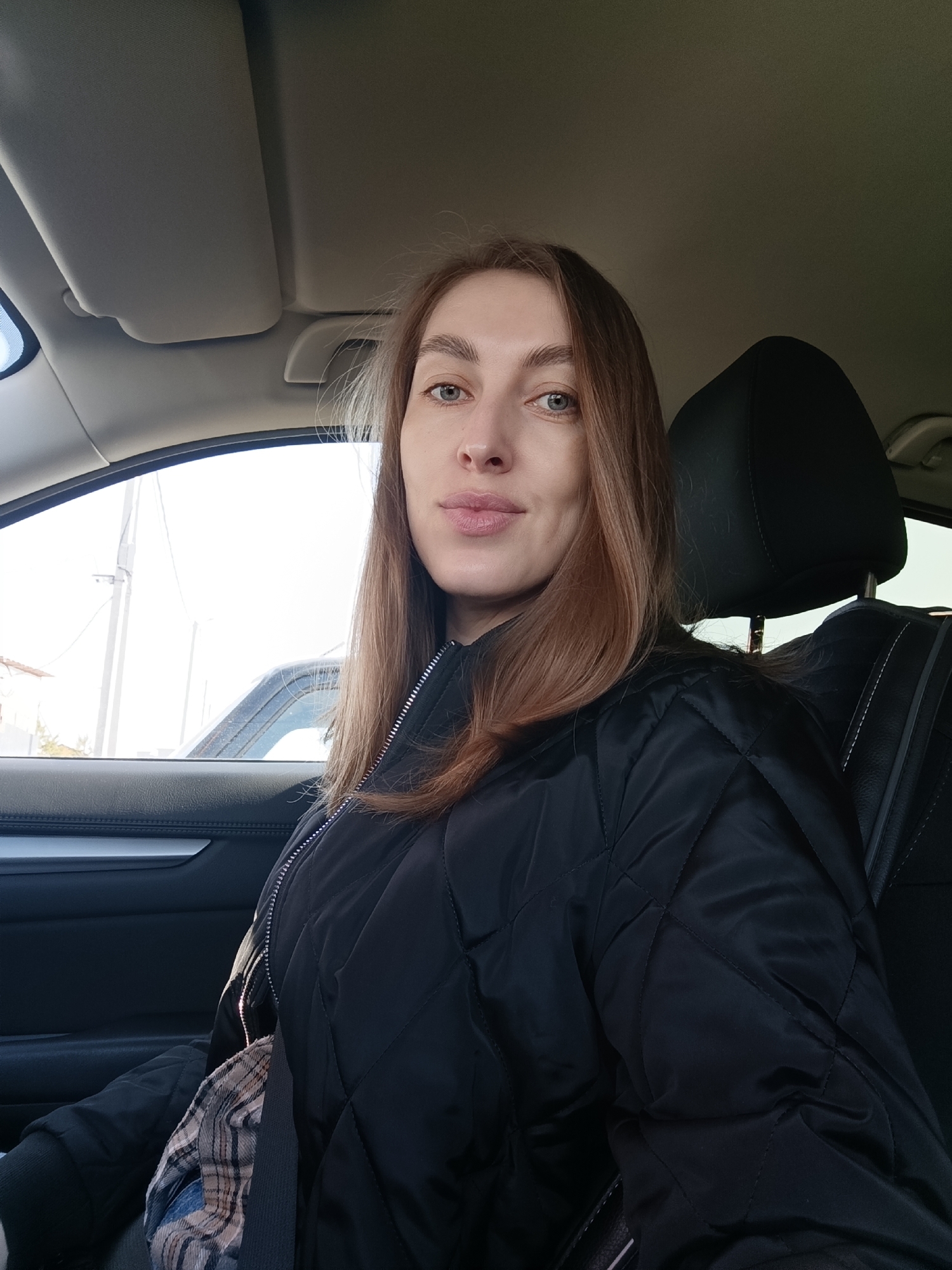 A woman is sitting in the driver's seat of a car. She is wearing a black quilted jacket and a seatbelt. The car is parked, and the view through the window shows a daytime urban scene with power lines and a building in the background. Without additional metadata, it's impossible to confirm the specific location as Chaikovsky, Russia.