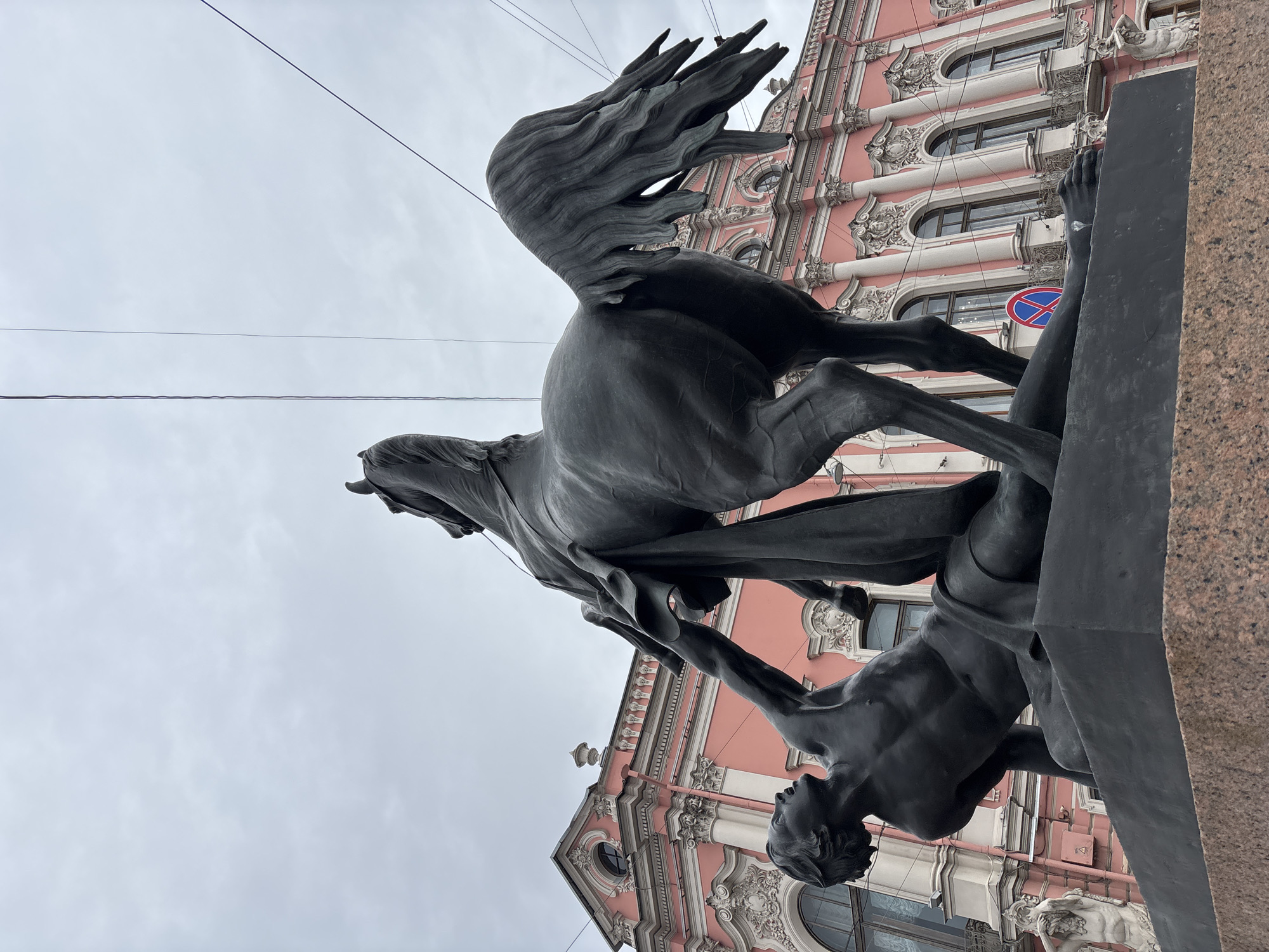 The image shows the equestrian statue of Peter von Klodt's "The Horse Tamers" on the Anichkov Bridge in Saint Petersburg, Russia. The statue features a nude man struggling to control a rearing horse. In the background is a pink building, likely a historic structure typical of Saint Petersburg's architecture. A road sign is visible on the building. The sky is overcast.