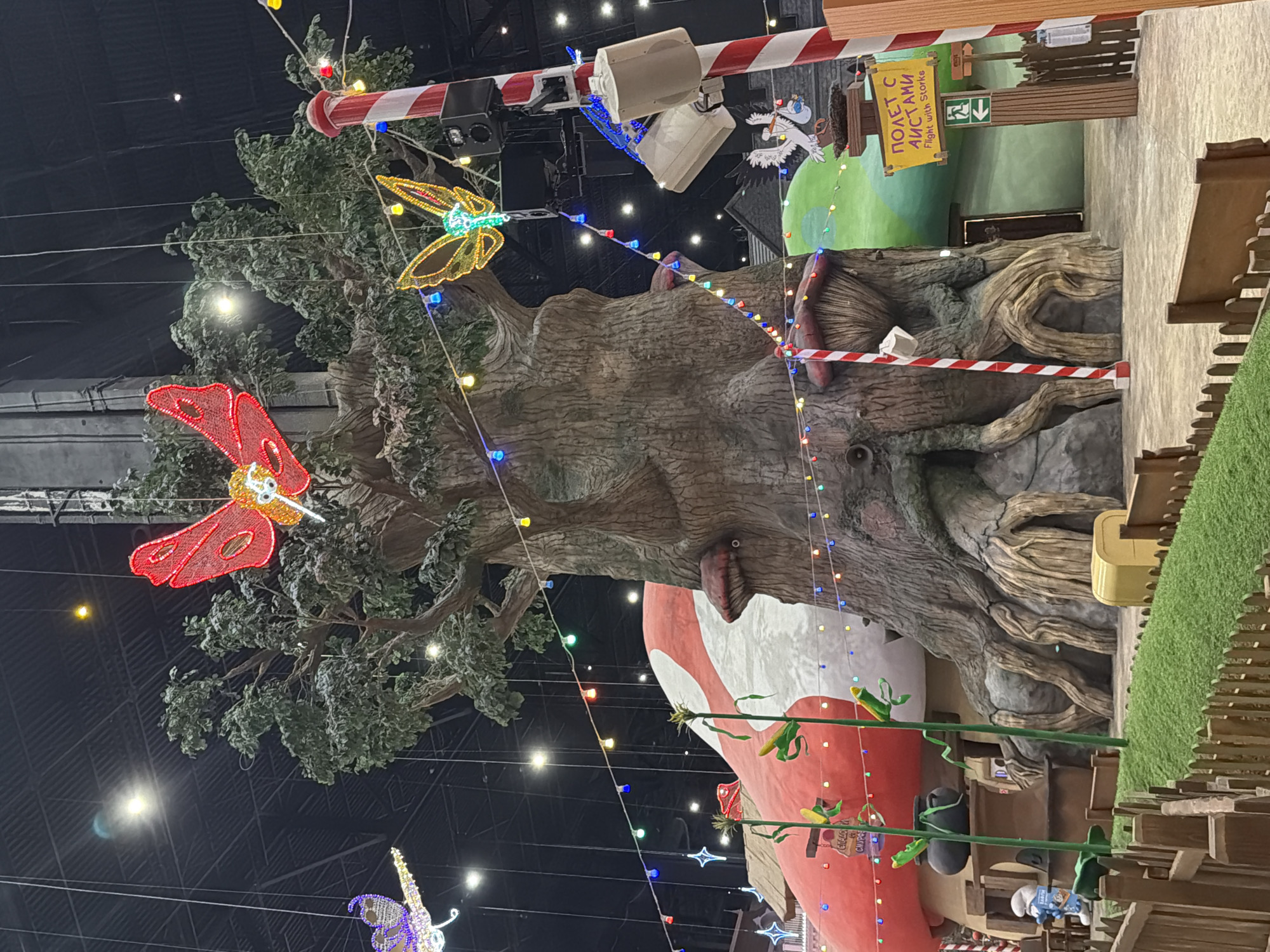 The image depicts an indoor whimsical environment featuring a large, stylized tree as the central element. The tree has exposed, gnarled roots and is adorned with numerous colorful LED lights strung across its branches and surrounding structures. Two large, illuminated butterfly decorations, one red and one yellow, are suspended from above. To the left of the tree is a giant red and white mushroom-shaped building. To the right, a red and white striped pole with speakers and lights is visible, and a sign with Russian text and an English translation reading "Flight with Storks" is mounted on a wooden post. A stork figurine carrying a baby is perched on top of a wooden structure. The environment appears to be a themed attraction or play area, with artificial grass, wooden fences, and other whimsical structures like corn stalks with Smurf-like figures. The ceiling above is dark, with a grid-like structure and various lights. The overall impression is one of a fantastical, illuminated indoor space.