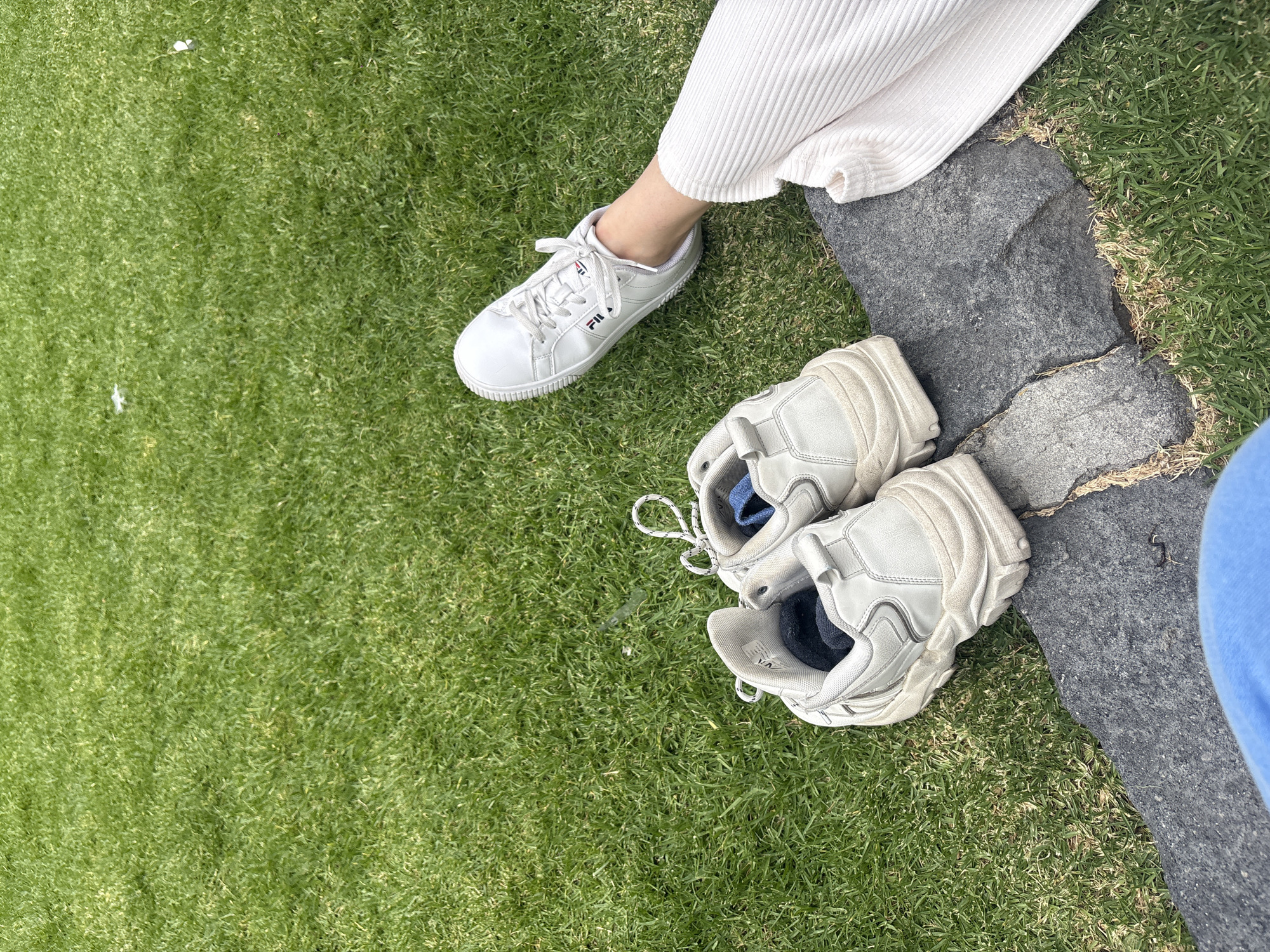 A high-angle medium shot depicts a person seated on a stone surface adjacent to grass. The individual is wearing a pair of white ribbed pants and has one white Fila sneaker on their left foot. Beside the stone, on the grass, there is a pair of white, chunky sneakers with laces untied. The location is Santa Ana Acozautla, Mexico.
