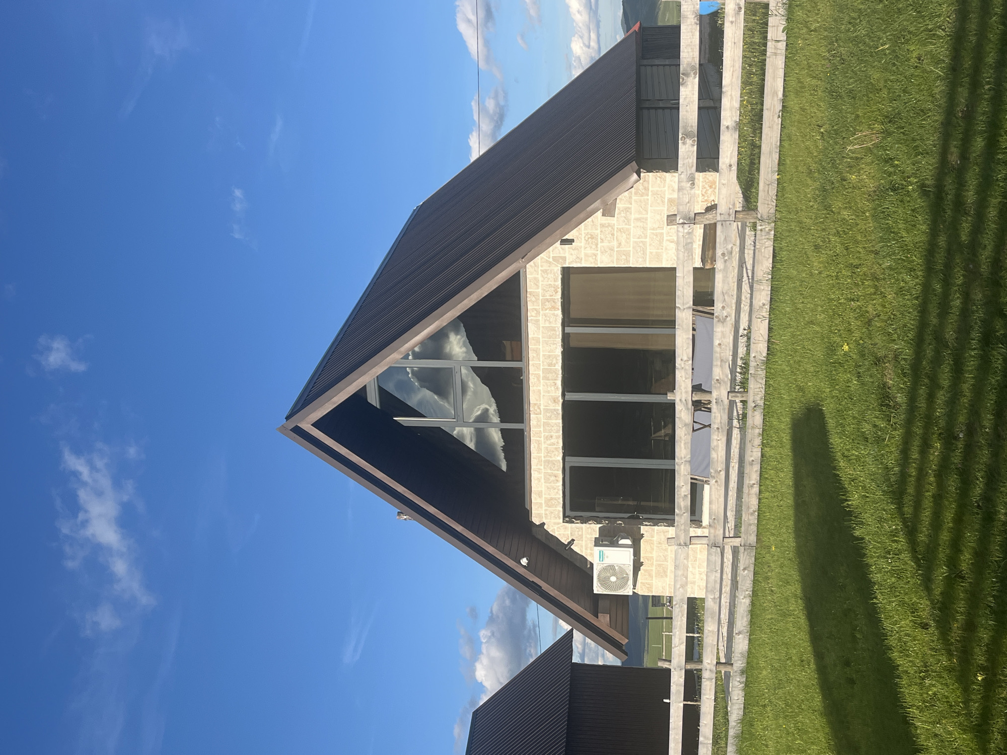 The image shows a modern A-frame house with a dark metal roof and light-colored stone exterior. The house has large windows, including a prominent triangular window at the apex of the roof that reflects the sky and clouds. An outdoor air conditioning unit is mounted on the wall to the left of the main entrance. A wooden fence runs across the foreground, separating the house from a grassy lawn. The background features rolling hills under a bright blue sky with scattered white clouds. To the left of the A-frame house, another smaller building with a dark metal roof is partially visible. The overall scene suggests a rural or semi-rural setting, possibly a vacation home or a newly constructed residence in the area of Virak, Montenegro. There are no people or animals visible in the image.