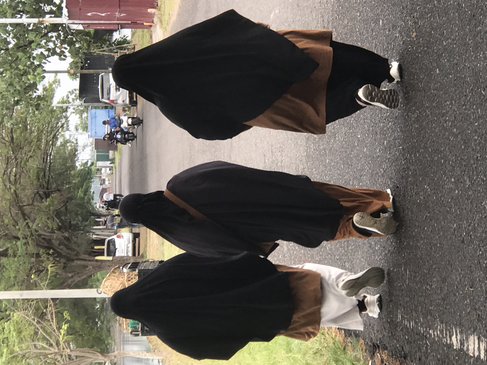 Three individuals wearing full black niqabs with visible ankle-length dresses in brown, white, and brown respectively, and sneakers, are walking on an asphalt road. The location appears to be a street in Lhokseumawe, Indonesia, evidenced by the local architecture and road conditions. In the background, there are cars, motorcycles, trees, and buildings. The road surface shows signs of wear. The sky is overcast, indicating the time of day is during daylight hours.