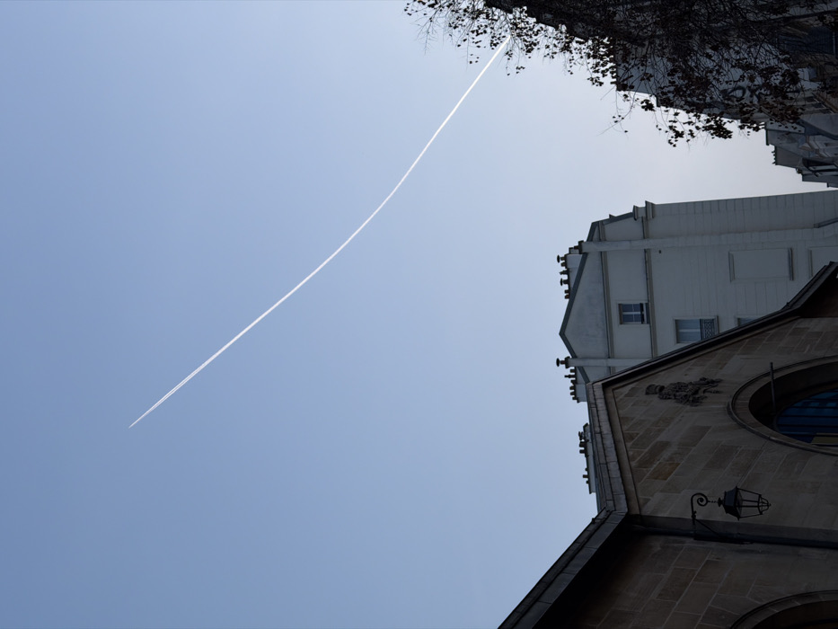 The image displays a clear, light blue sky traversed diagonally by a prominent, white aircraft contrail from the upper left to the lower right. Below the sky, an urban environment consistent with Paris, France, is depicted.
In the lower left, the upper portion of a large building constructed from light brown/tan stone blocks is visible. This structure features a pitched roofline, a decorative stone crest or coat of arms affixed to its facade, and a large arched opening below the crest. An ornate, black metal lantern hangs from the stone wall to the left of the crest.
Behind and to the right of this stone building, a taller, multi-story building with a light-colored (appears off-white or light grey) facade stands, displaying architectural elements characteristic of Haussmannian design, including multiple dark chimneys along its roofline. Several windows are visible on its upper floors.
Further to the right, a section of a dark, bare-branched tree, possibly deciduous, partially obscures the sky, the contrail, and another building with a pitched roof and visible chimneys.
No people, discernible actions, or interactions are visible in the frame. The lighting suggests daytime conditions, with sunlight illuminating parts of the scene, particularly affecting the contrail and the right side of the buildings.