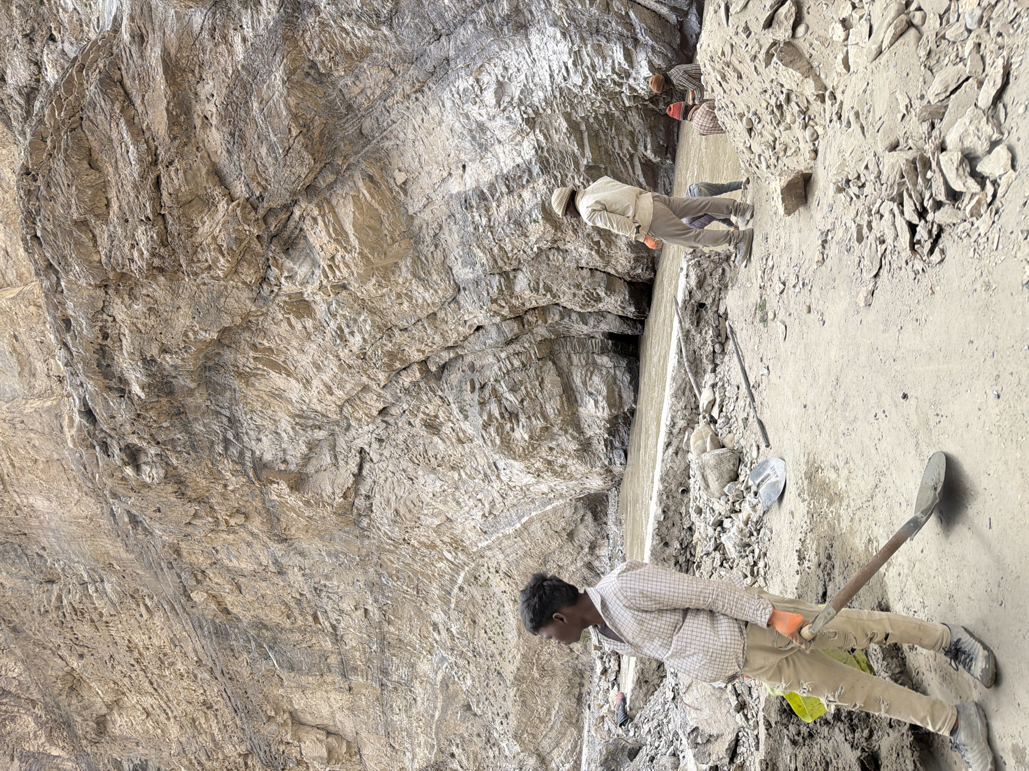In Stongde, India, a group of men are working in a rocky, mountainous area beside a muddy river. The terrain is rough, with loose stones and debris scattered everywhere. A large, imposing cliff face dominates the background, its striated layers of rock suggesting geological history. In the foreground, a young man wearing a plaid shirt, ripped trousers, and orange gloves stands with a shovel, looking off to the left. Further back, another man in a light-colored jacket and cap stands near the riverbank, facing away from the camera. Two other individuals, one with a red hat, are seated or crouched by the river. The overall impression is one of manual labor in a rugged natural environment.