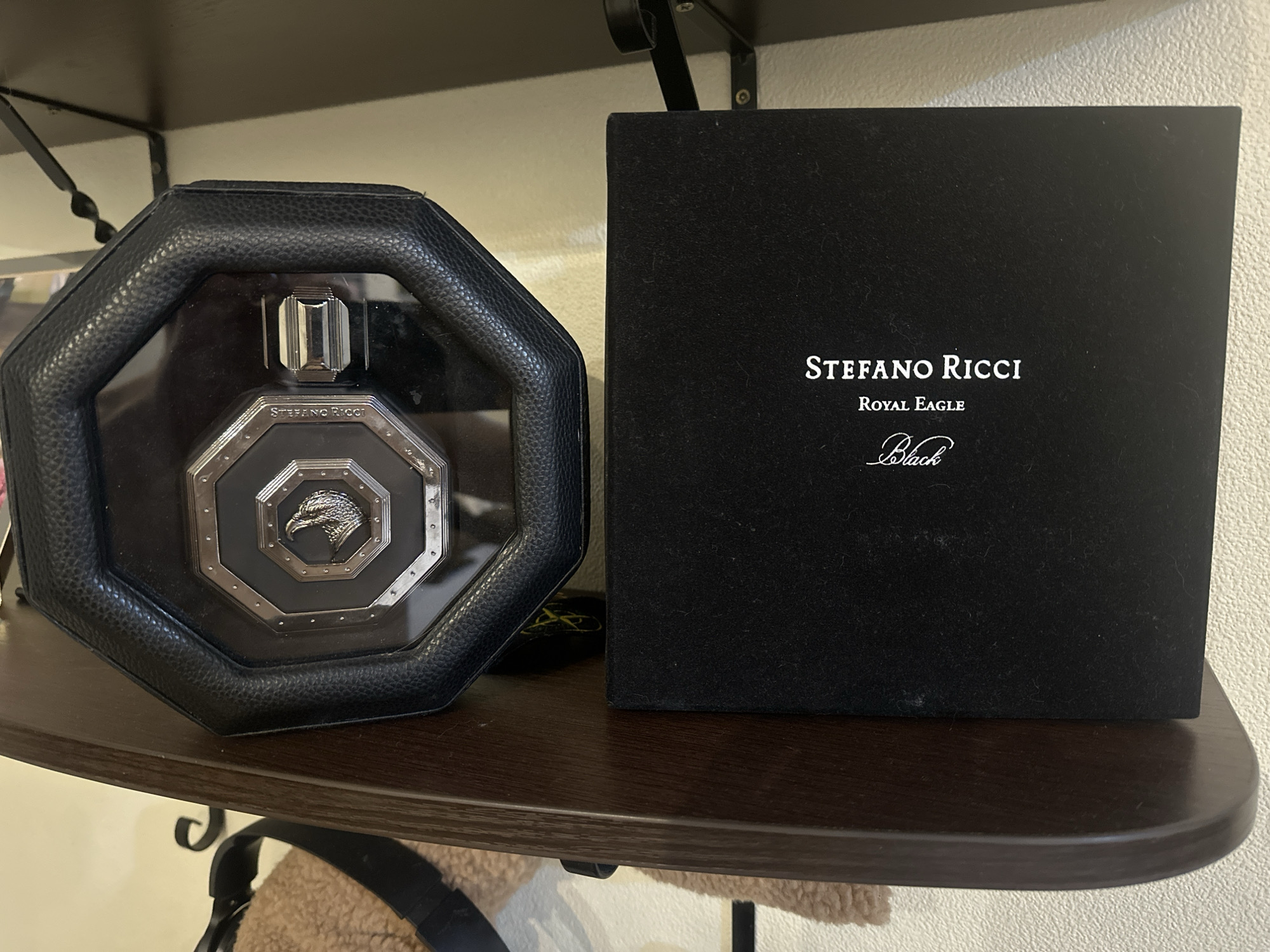 The image shows a black box with "STEFANO RICCI ROYAL EAGLE Black" printed on it in white text, and an octagonal black leather case containing a perfume bottle with a silver cap and an eagle emblem on the front, placed on a wooden shelf.