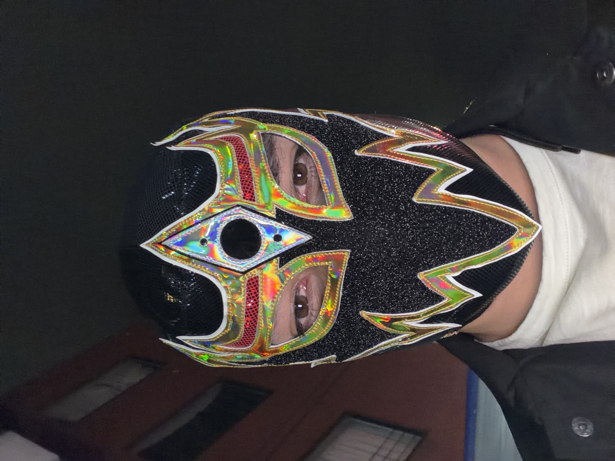 A person wearing a black lucha libre mask with iridescent and gold trim is facing the camera. The mask has an eye shape with a hole in the center. The person's eyes are visible through the eye shapes of the mask. The person is wearing a white shirt and a black jacket. The background is dark, with a glimpse of a brick building with windows in the lower left corner. Based on the style of architecture, this could possibly be a location in the city of Puebla, Mexico.