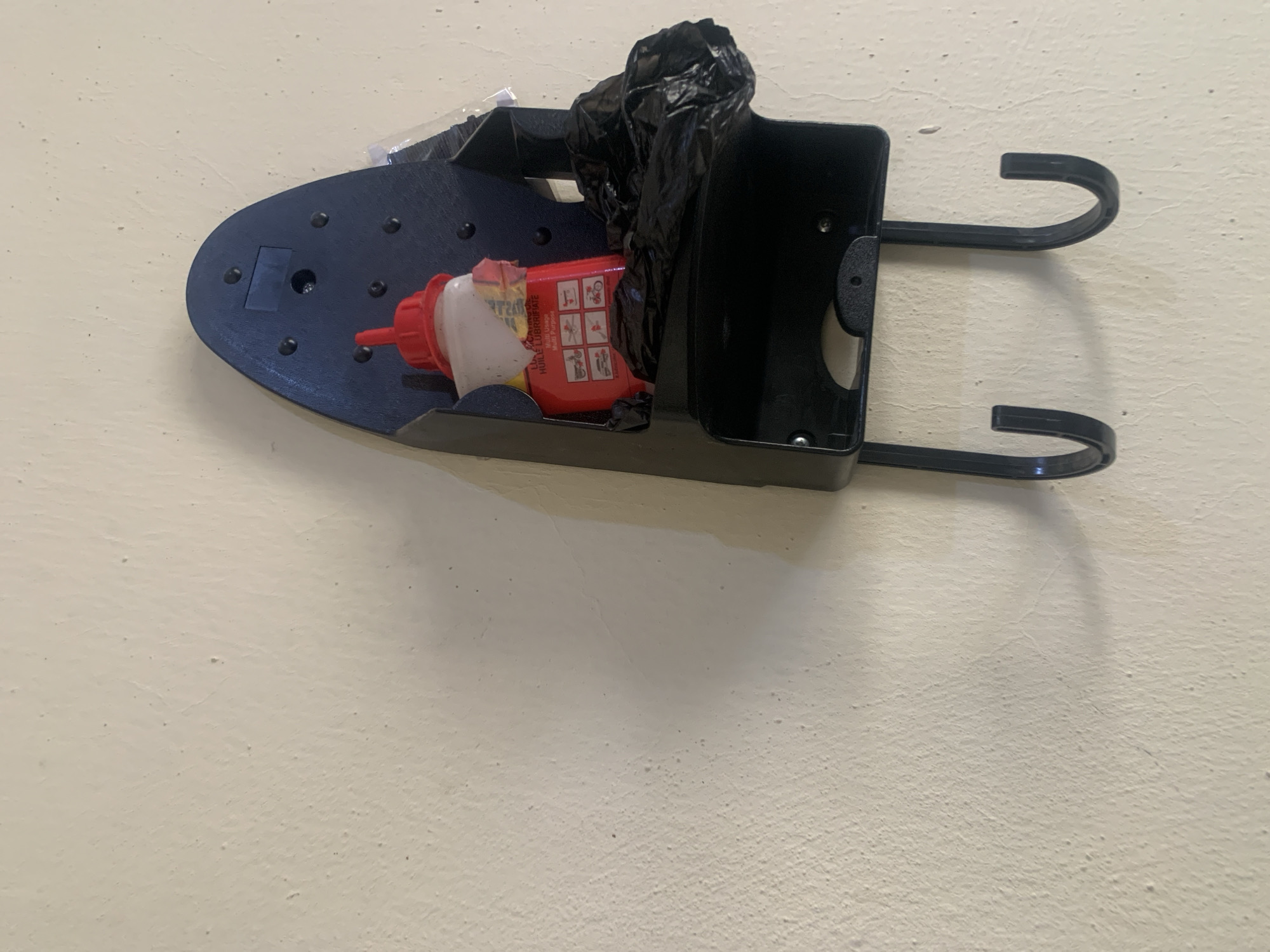 A wall-mounted ironing board holder, dark blue in color, is affixed to a cream-colored textured wall. The holder has a narrow, curved top section and a wider, rectangular section below with two prominent black hooks extending downwards. Within the rectangular section, a red-capped white bottle of lubricant is placed, alongside a crumpled black plastic bag. The bottle has visible labels with text and images, including depictions of a motorcycle, car, and bicycle. The lighting suggests an indoor environment, possibly a storage area or utility room. The location of Jauro Yinu, Nigeria is not discernible from the visual information provided in the image.