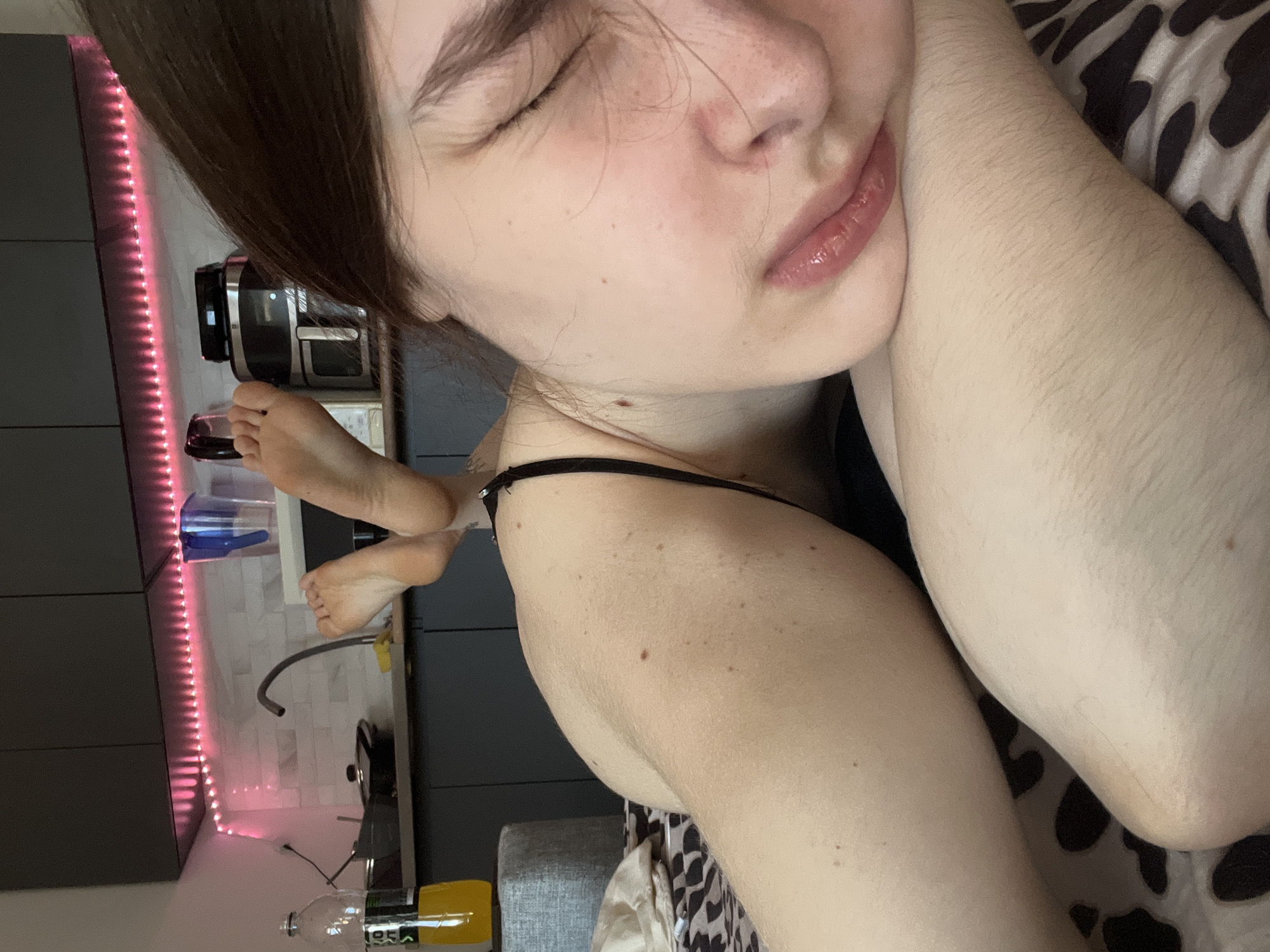 A young woman with closed eyes is lying down, her face visible, and her right arm is bent and positioned near her head. She is wearing a black strap tank top. Her feet are up and appear to be resting on a kitchen counter. The counter is a gray color and has a stainless steel toaster oven, and a blue plastic container on it. Underneath the counter are a stainless steel faucet, a black cooking pot, and a yellow bottle of liquid. The scene is lit by pink LED strip lights. A gray ottoman and bedding with a black and white animal print are also visible. It is impossible to determine the location in Saint Petersburg, Russia.
