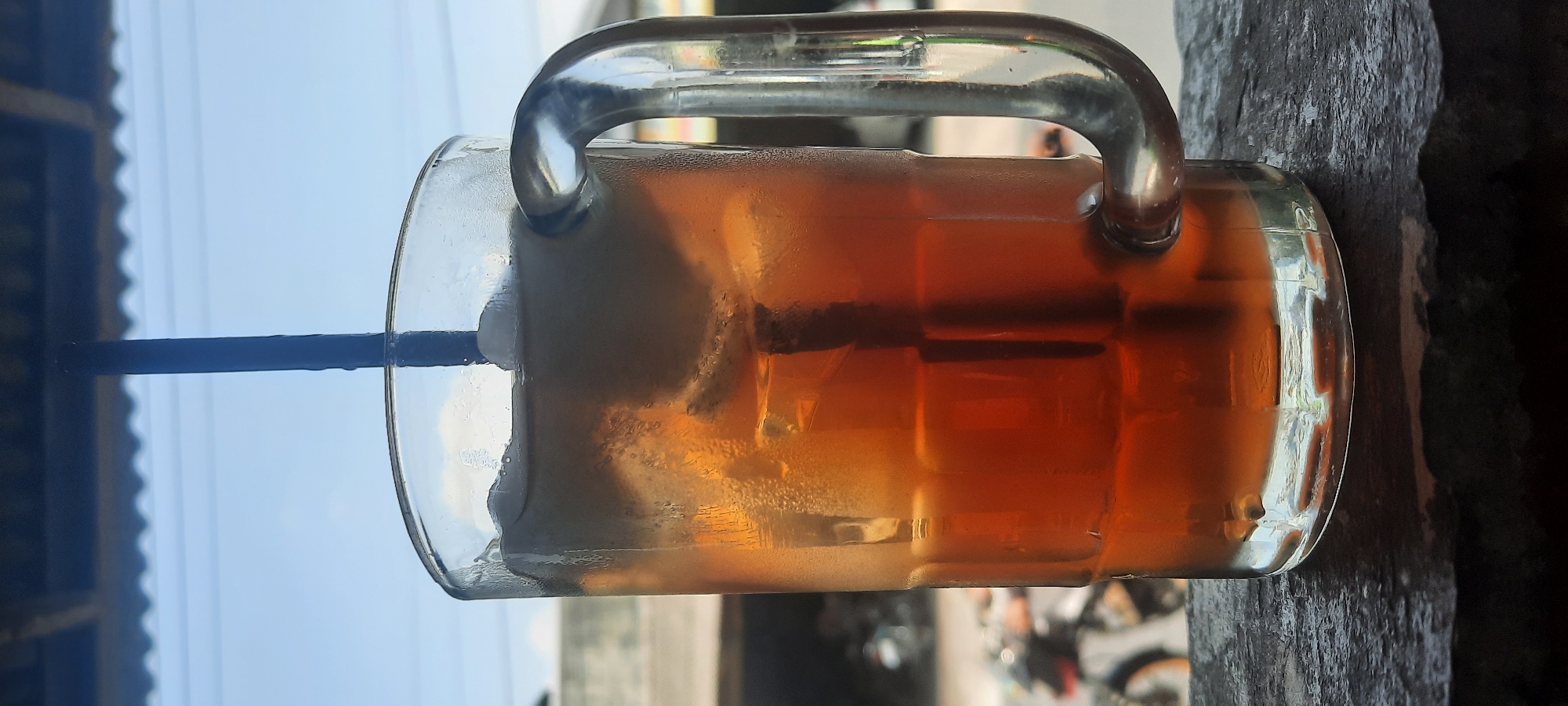 A tall glass mug filled with iced tea sits on a dark, textured surface. The mug has a thick glass handle and is filled with a reddish-brown liquid and several ice cubes. A dark blue straw extends from the liquid to the top of the mug. The background is blurred, showing a bright sky with some clouds, a corrugated roof structure, and the indistinct shapes of buildings and a motorcycle. The image appears to be taken outdoors, possibly on a patio or balcony, with a shallow depth of field that focuses on the drink. The setting is identified as Selopuro, Indonesia.
