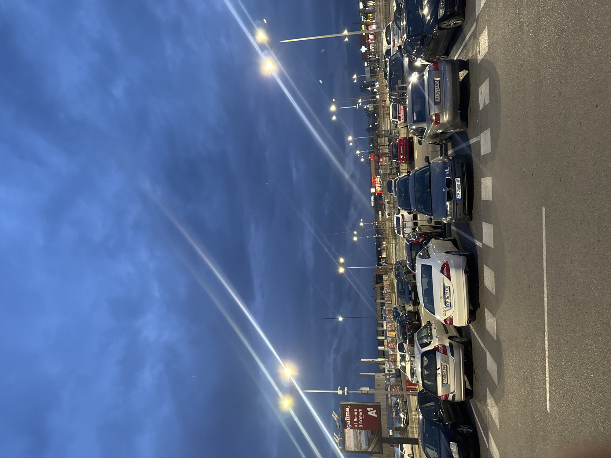 The image shows a parking lot at night, likely in a shopping mall or similar commercial area. The sky is a dark blue with some clouds. Numerous cars are parked in rows, with license plates visible on some of them. The license plates appear to be Bulgarian. Streetlights illuminate the area, and a large billboard is visible in the foreground, advertising the A1 telecommunications company and XOPark, which is a large outlet store in Sofia, Bulgaria. The vehicles are parked adjacent to a building with visible retail operations. Based on these elements, the location is likely the parking area for XOPark in Sofia, Bulgaria.