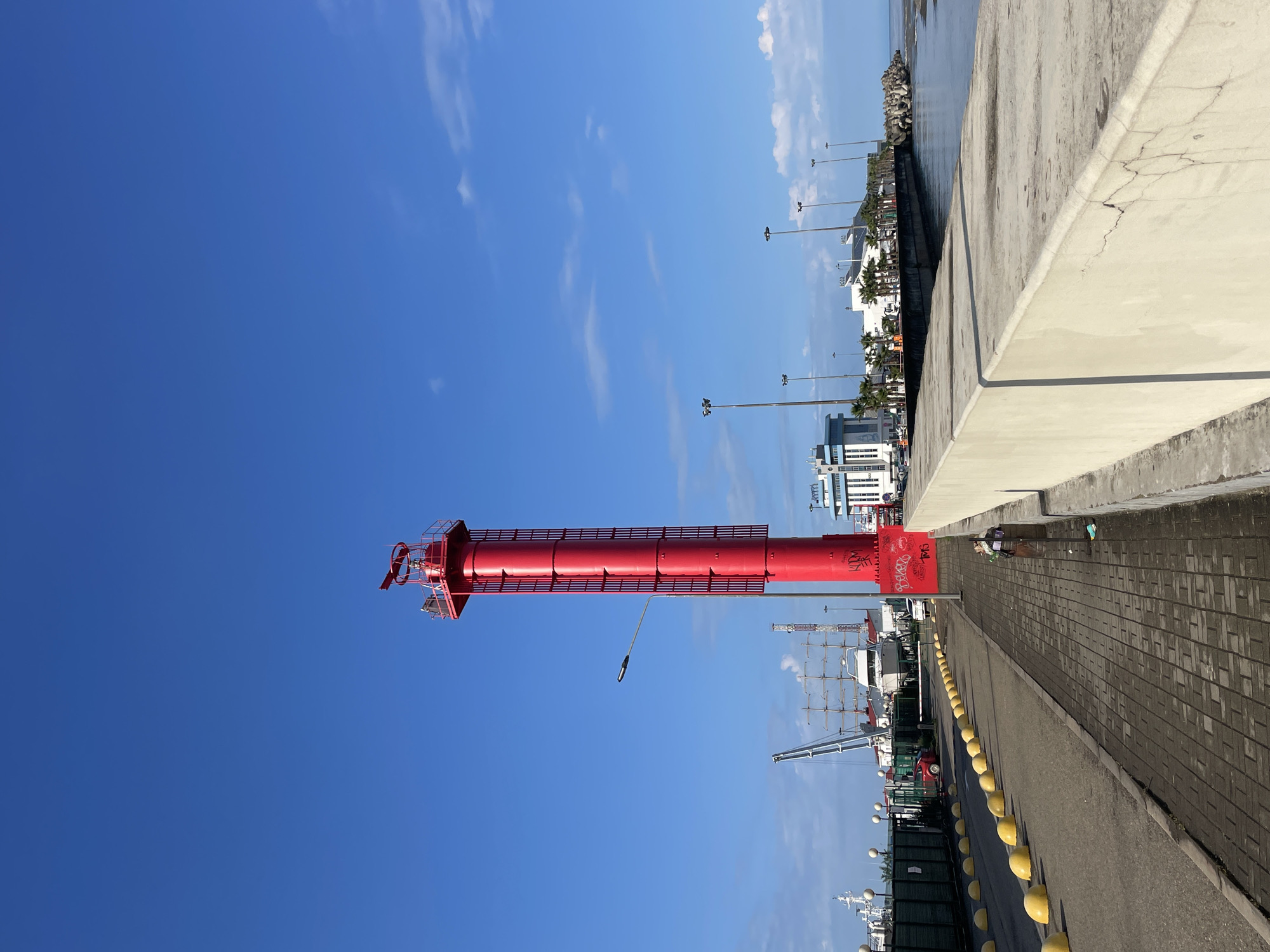 The image shows a tall, red lighthouse standing next to a paved walkway with yellow bollards, with a clear blue sky above. The lighthouse appears to be in a harbor area, with various boats and cranes visible in the background. Buildings and streetlights can also be seen further in the background. The presence of palm trees suggests a coastal environment. The graffiti on the base of the lighthouse indicates some urban activity. Given the prompt, the location is likely the city of Sochi, Russia.