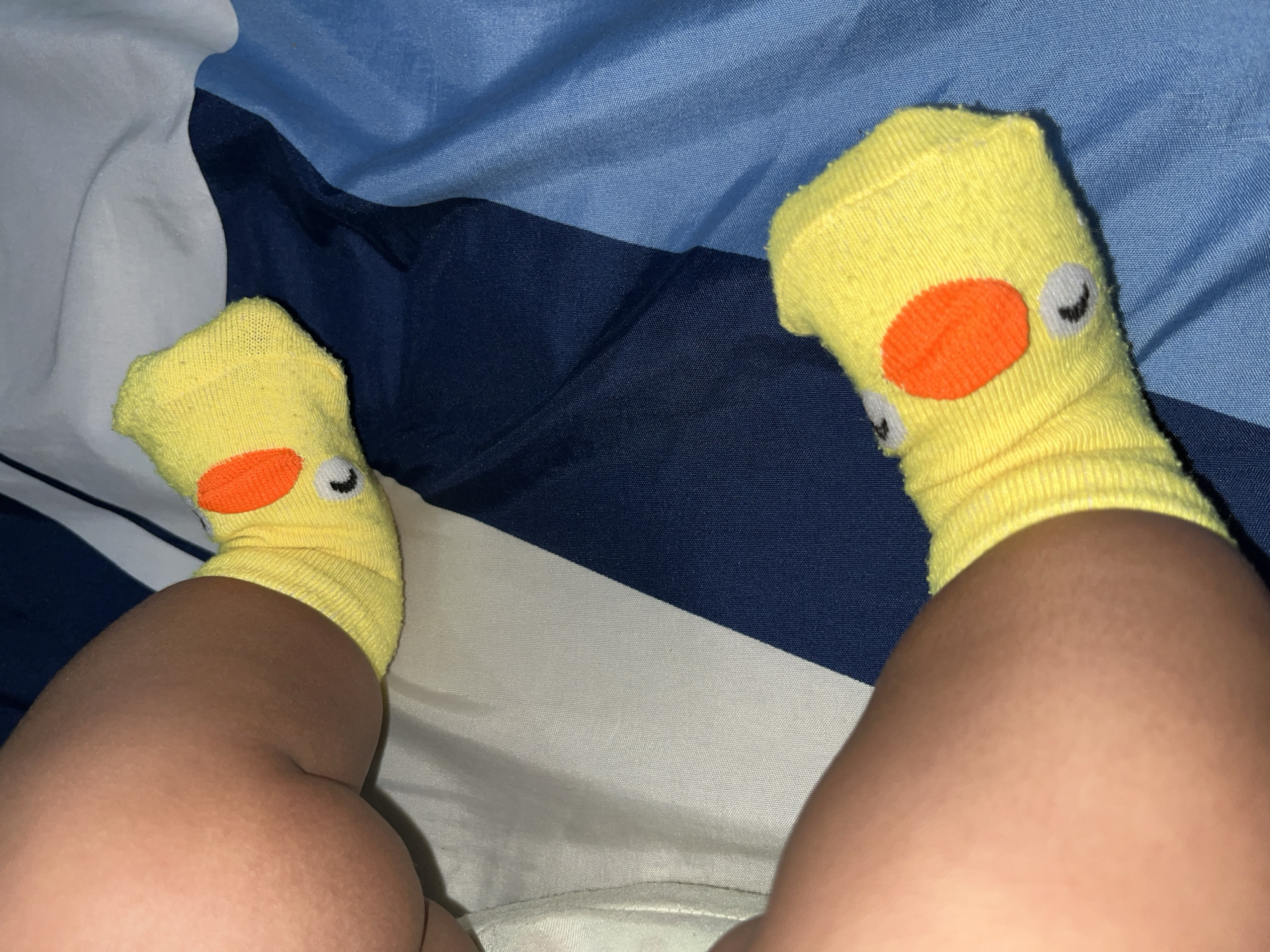 The image shows two legs wearing yellow socks with a duck face design (orange beak and closed eye). The legs are resting on a bed with blue and white striped bedding. The scene is indoors, likely in a private residence. The location in the city of Lhokseumawe, Indonesia cannot be determined from the image.