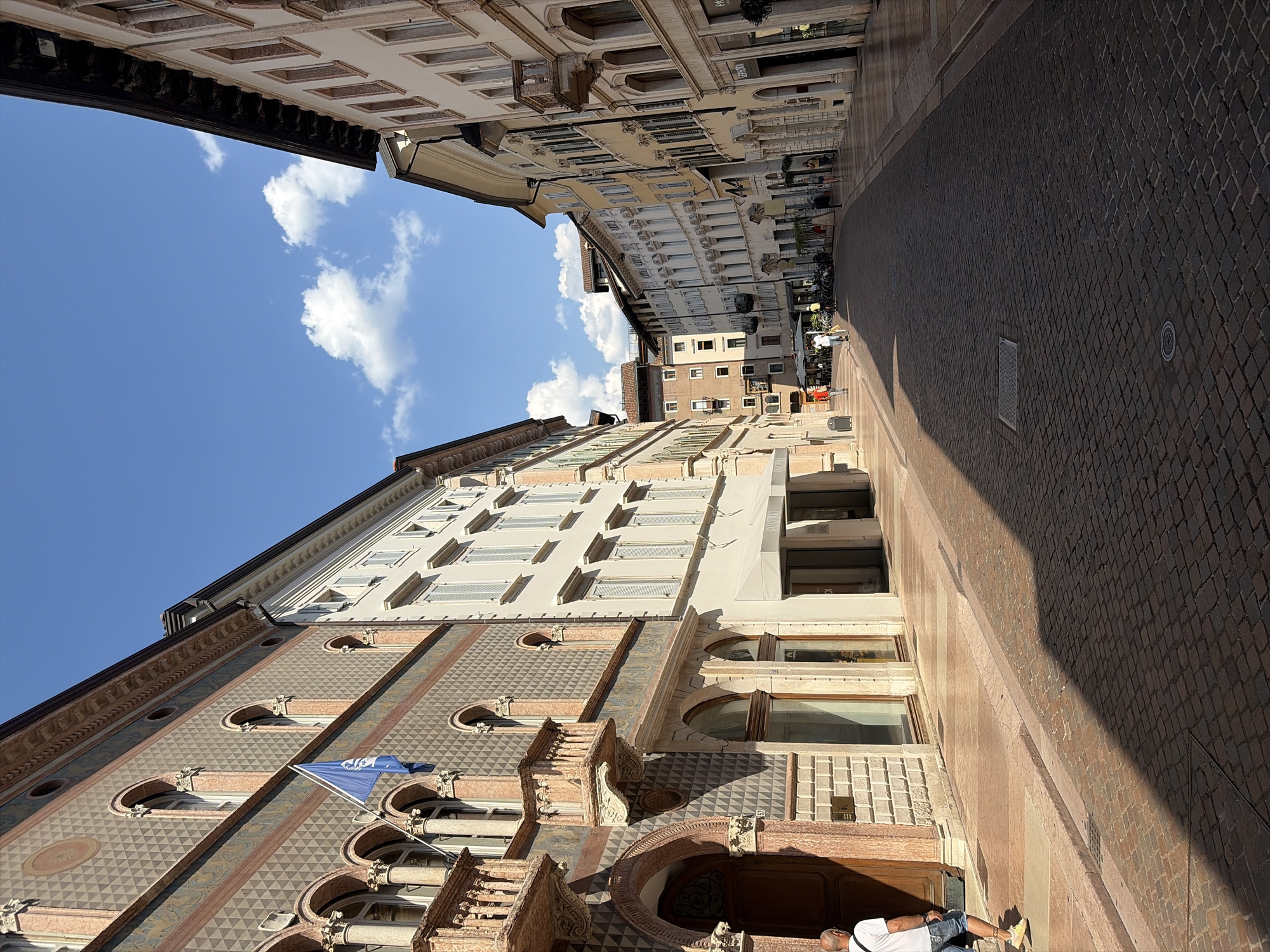 The image shows a view looking down a cobblestone street in the historic center of Trento, Italy. The street is lined with ornate, multi-story buildings with arched windows and balconies. The architecture suggests a historic European city, and the presence of a blue flag with a crest indicates a potential official building. On the left, a man wearing a white t-shirt and denim shorts walks away from the camera. The street is partially shaded by the buildings, with sunlight illuminating sections of the cobblestones and the facades. The sky above is blue with scattered white clouds. Several shops with awnings are visible along the street. The overall scene is one of a sunny day in a picturesque urban environment.