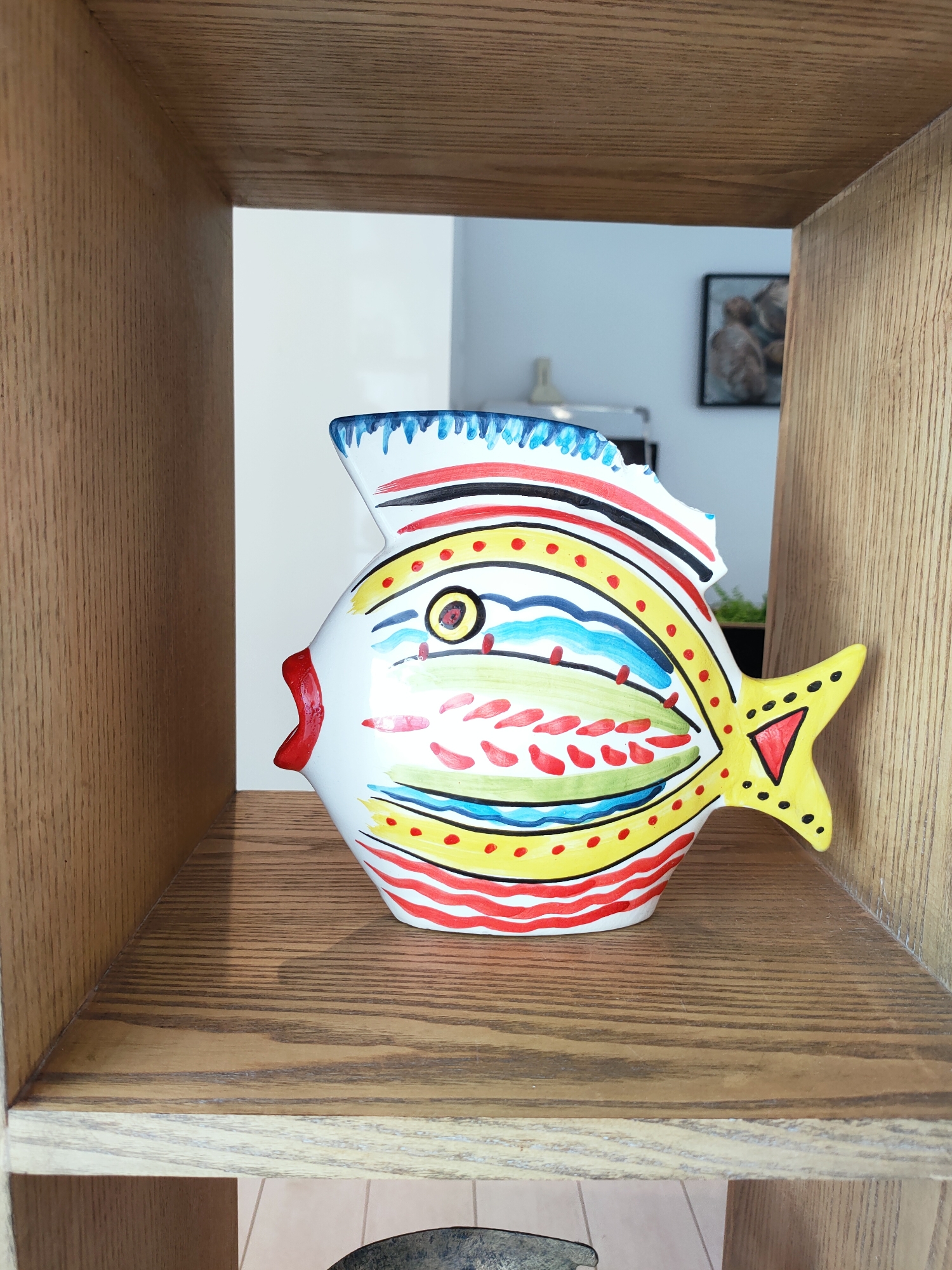 A colorful ceramic fish figurine sits on a wooden shelf in a room. The fish is white with a red mouth, a yellow tail with a red triangle, and is decorated with various patterns in red, yellow, blue, and green. The shelf is made of light-colored wood with visible grain. In the background, a wall is visible, and a framed picture hangs on it. The overall scene suggests an indoor setting, possibly a home or a shop. The location is identified as the city of Tunis, Tunisia.