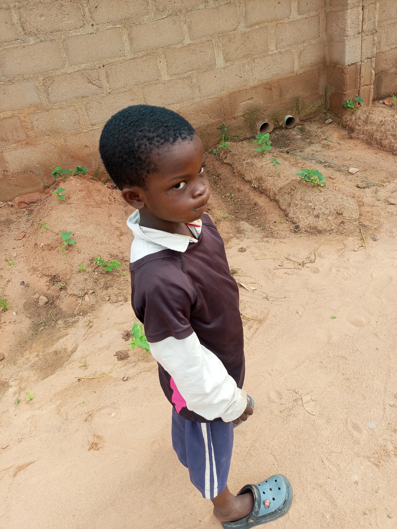 A young boy stands in an outdoor environment. He is looking towards the camera with a slight tilt of his head. The boy is wearing a dark t-shirt with white sleeves, dark blue shorts, and a grey croc-style sandal on his left foot. He is standing on sandy ground, and behind him is a light-colored brick wall. There are some small green plants growing in the sandy area and near the wall. Two drainage pipes are visible protruding from the wall. The scene appears to be in an unpaved area, possibly a yard or undeveloped lot, in Tempure, Nigeria.