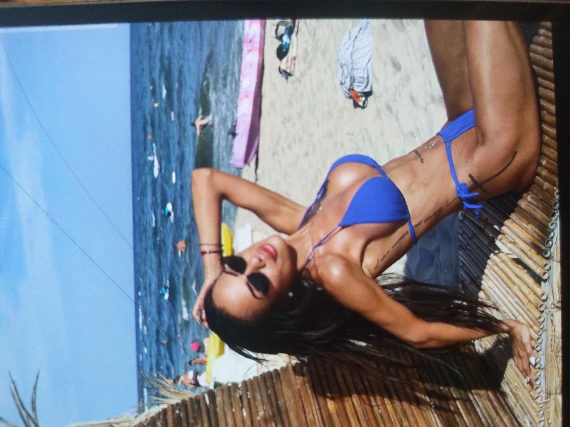 The image shows a woman in a blue bikini lying on a woven surface, likely a beach mat or lounge chair. She is wearing sunglasses and her long dark hair is spread out. In the background is a beach with people, surfboards, and the sea. Given the prompt, it is implied this is taken on the beach in Tsarevo, Bulgaria.