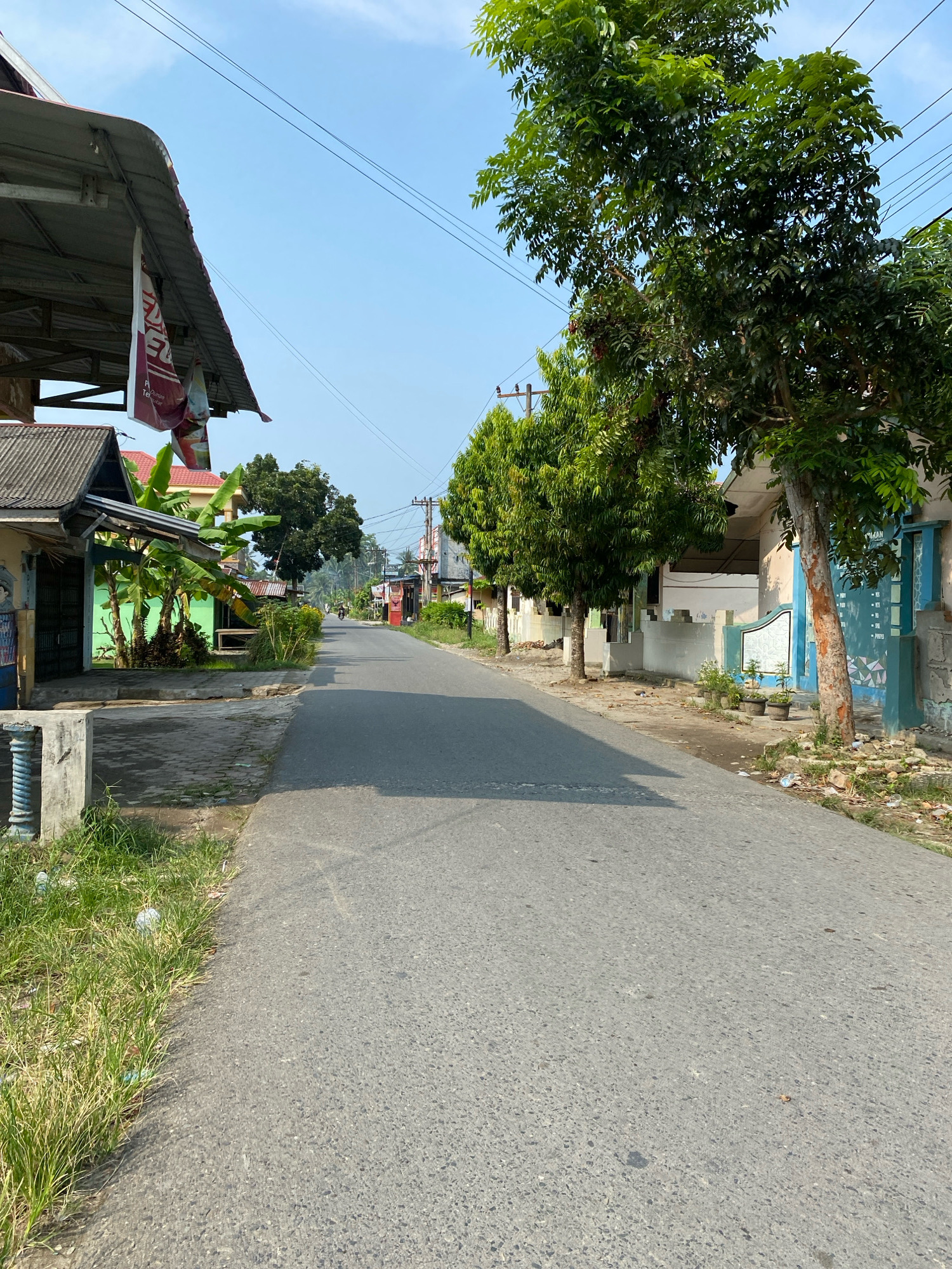 The image depicts a straight, paved road in Karang Gading, Indonesia, likely a residential area, under a clear blue sky. On the left side of the road, there are several low-lying buildings with corrugated metal roofs, some adorned with banners. Lush green banana trees are visible behind the structures. The right side of the road features houses with light-colored walls and blue accents, lined by a row of trees with dense green foliage. A single figure on a motorcycle is seen in the distance, heading away from the camera. The road is mostly empty, with shadows cast across its surface from the surrounding trees and buildings, indicating it is likely mid-morning or afternoon. The environment appears somewhat unkempt, with patches of grass and some scattered debris along the roadside.