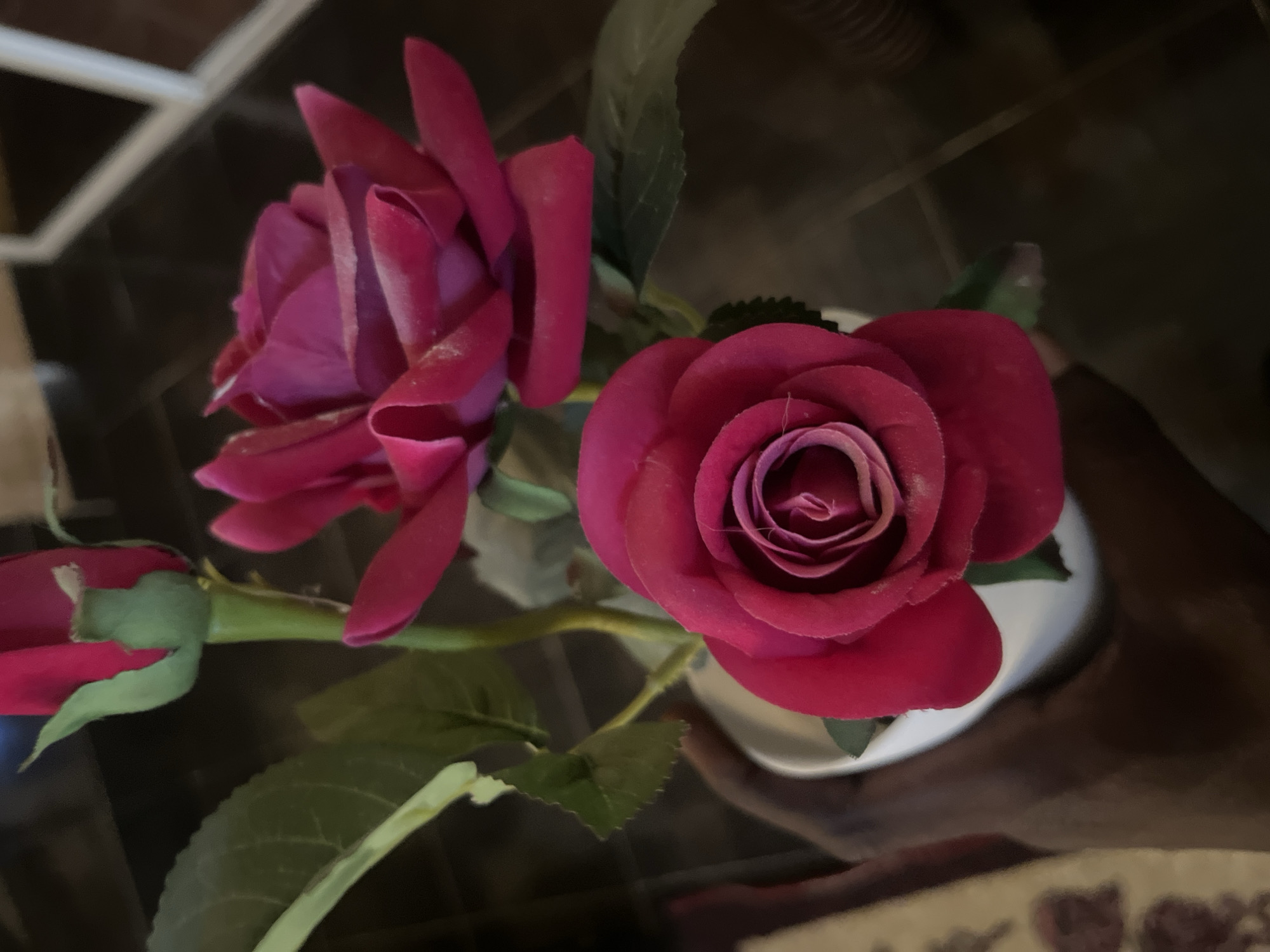 A close-up, waist-level shot shows three artificial pink roses in a white vase. A dark-skinned hand is holding the vase, with the fingers wrapped around the lower portion. The roses are vibrant pink, with delicate petals and some appearing slightly dusty. The background is dark and out of focus, with hints of wooden furniture or flooring. The scene appears to be indoors, potentially in a home or office setting. There is no indication of the location being in Barakin Akawo, Nigeria.