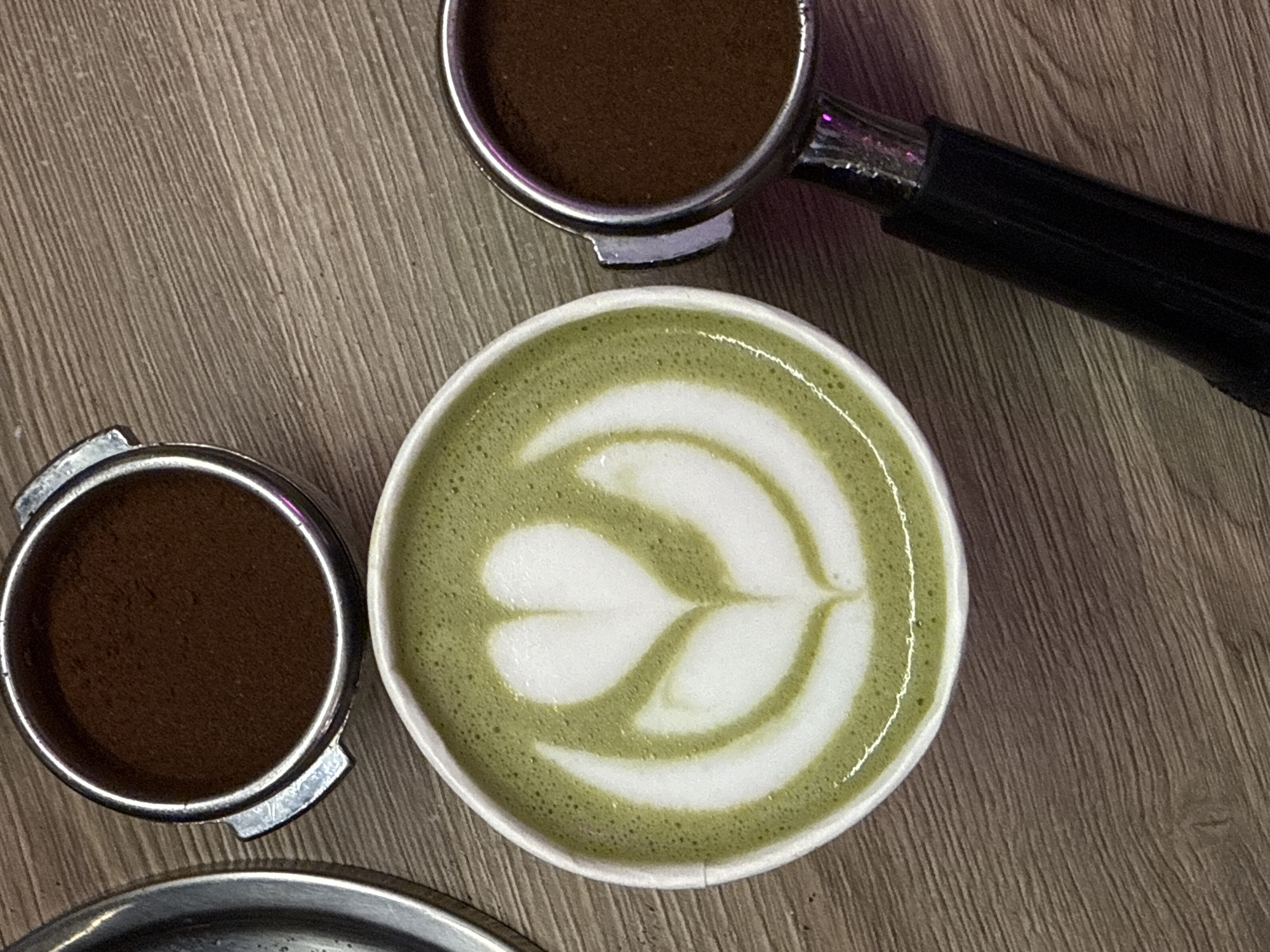 An overhead shot displays elements of a coffee preparation station, likely in Moscow, Russia. Dominating the center is a white ceramic cup containing a vibrant green matcha latte, adorned with intricate white latte art depicting a stylized heart or tulip design, indicative of skilled barista work. The latte's surface shows small, uniform bubbles in the green area, contrasting with the smooth white foam.
Two portafilters, typically used with espresso machines, are positioned around the cup. The upper right features a portafilter filled with dark brown, finely ground coffee, its metallic rim and a portion of its black handle visible. A subtle purple reflection is seen near the handle. Another similar portafilter, also holding ground coffee, is partially visible in the lower-left quadrant.
The objects are arranged on a light brown wooden surface, characterized by its linear grain pattern. A metallic tray or drip tray is partially visible at the very bottom edge of the frame. The scene suggests a moment during or after coffee and matcha preparation, possibly in a café or home setting. No text is visible in the image.